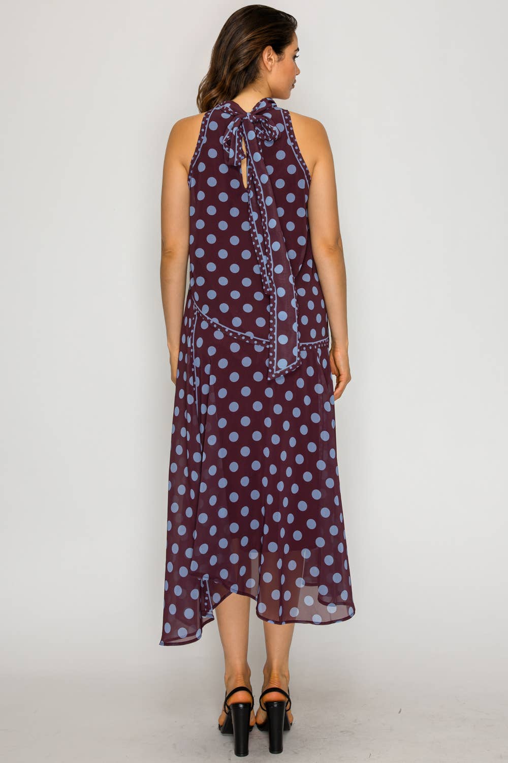 A woman viewed from the back wearing a sleeveless halter-neck midi dress with a purple and light blue polka dot pattern and a tie detail at the back neckline, paired with black high-heeled sandals against a plain light gray studio background.