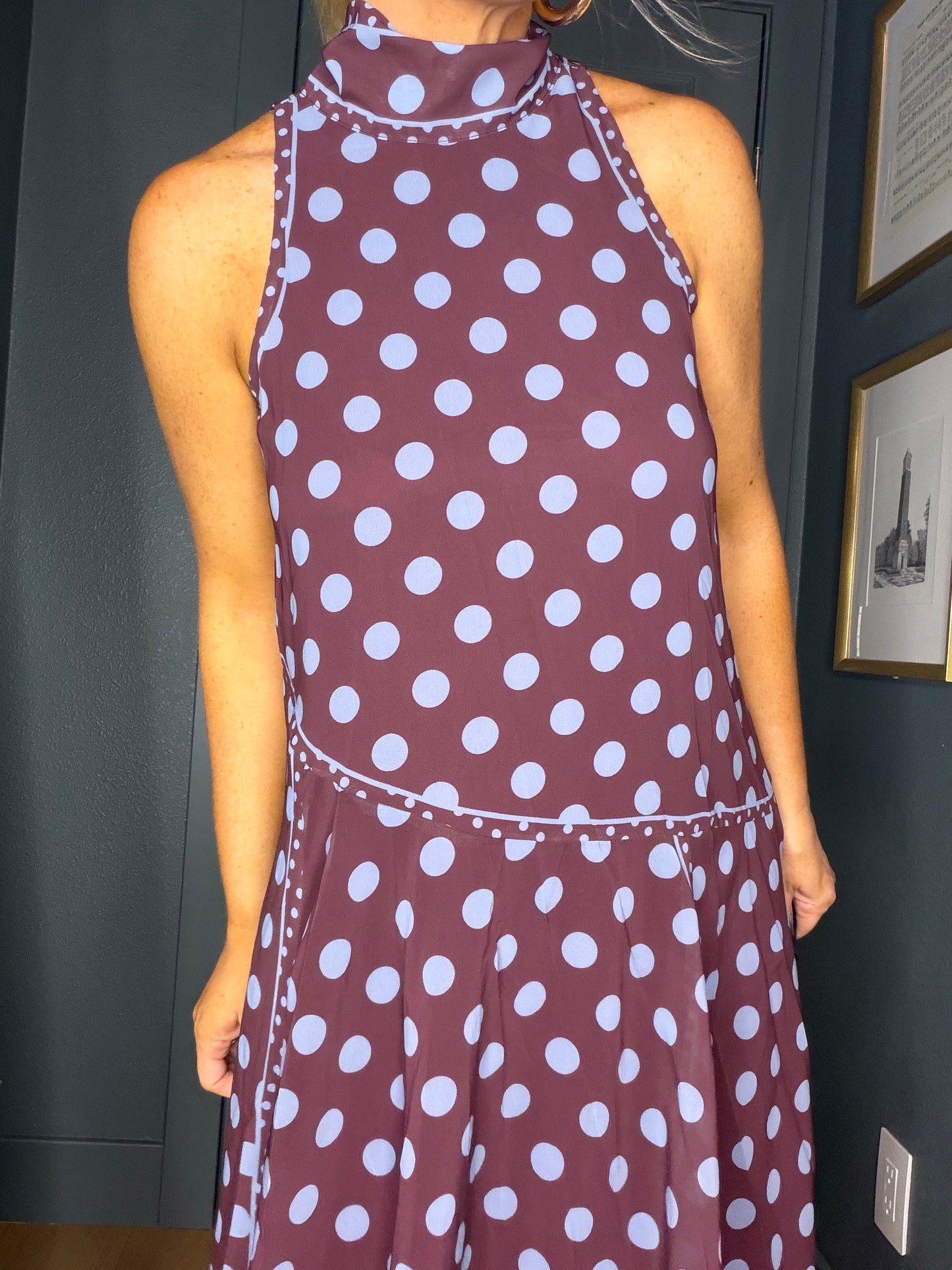 A sleeveless halter-neck dress with a dark plum base and white polka dots, shown on a model in an indoor setting.