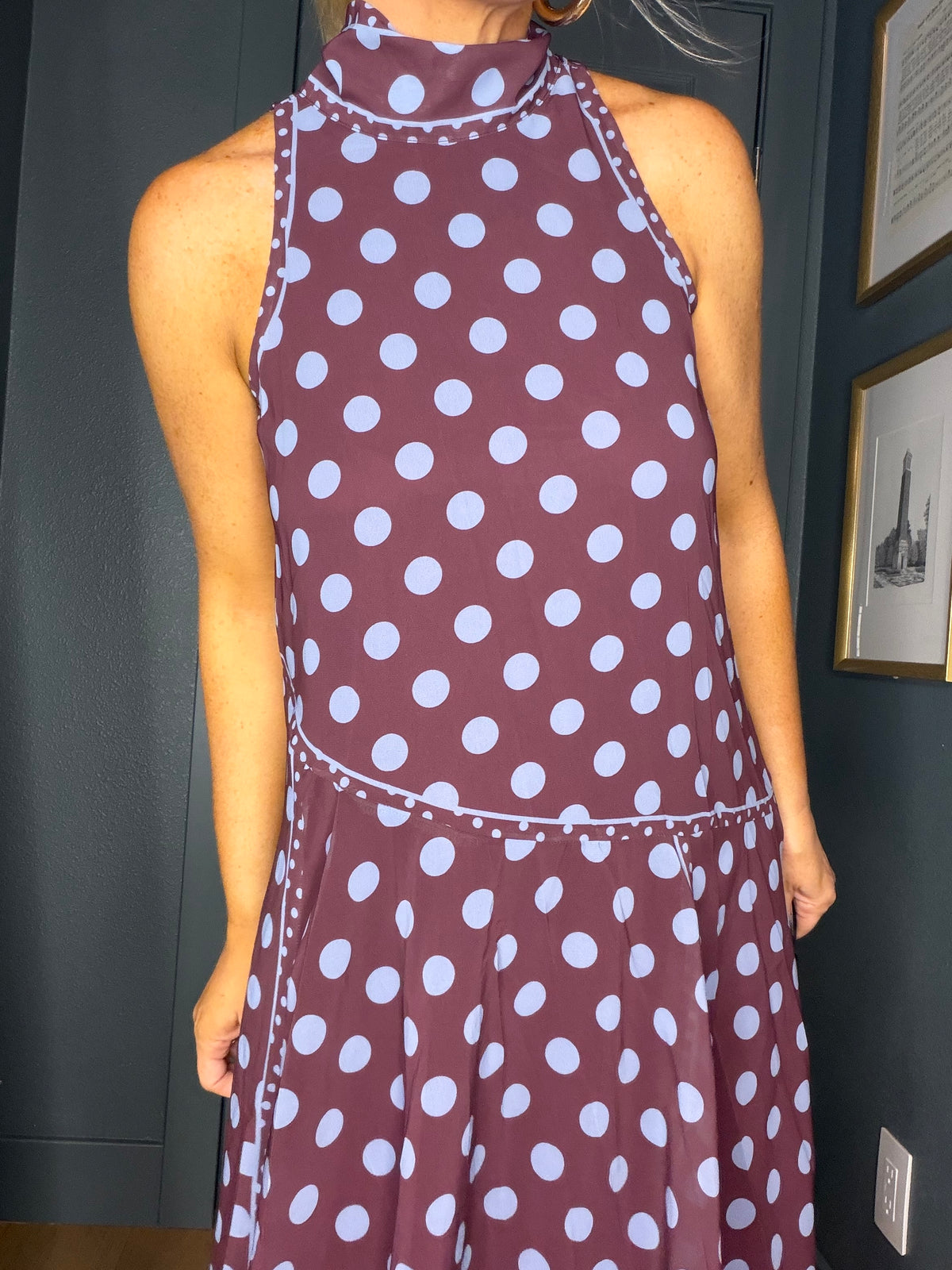 A sleeveless halter-neck dress with a dark plum base and white polka dots, shown on a model in an indoor setting.