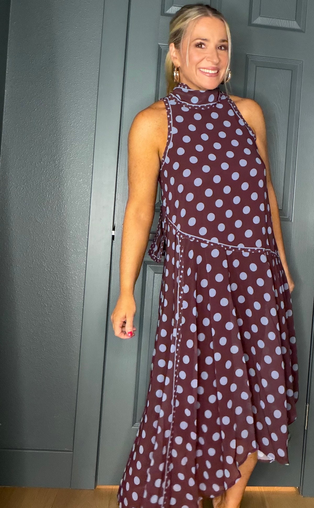A woman modeling a sleeveless high-neck midi dress with a brown base and light blue polka dots, standing in front of a dark gray paneled door.