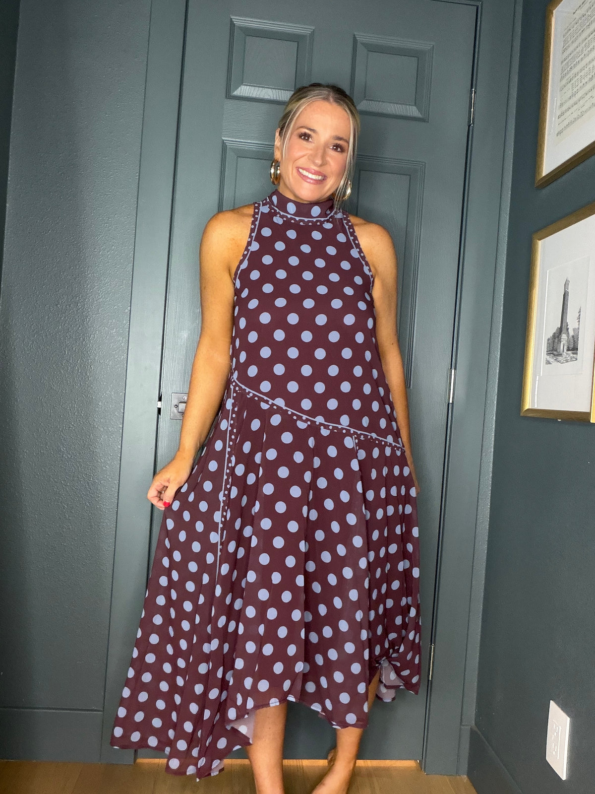 A woman wearing a sleeveless high-neck midi dress with a dark burgundy base and light blue polka dots, standing in front of a dark gray paneled door.