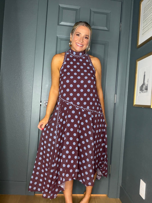 A woman wearing a sleeveless high-neck midi dress with a dark burgundy base and light blue polka dots, standing in front of a dark gray paneled door.