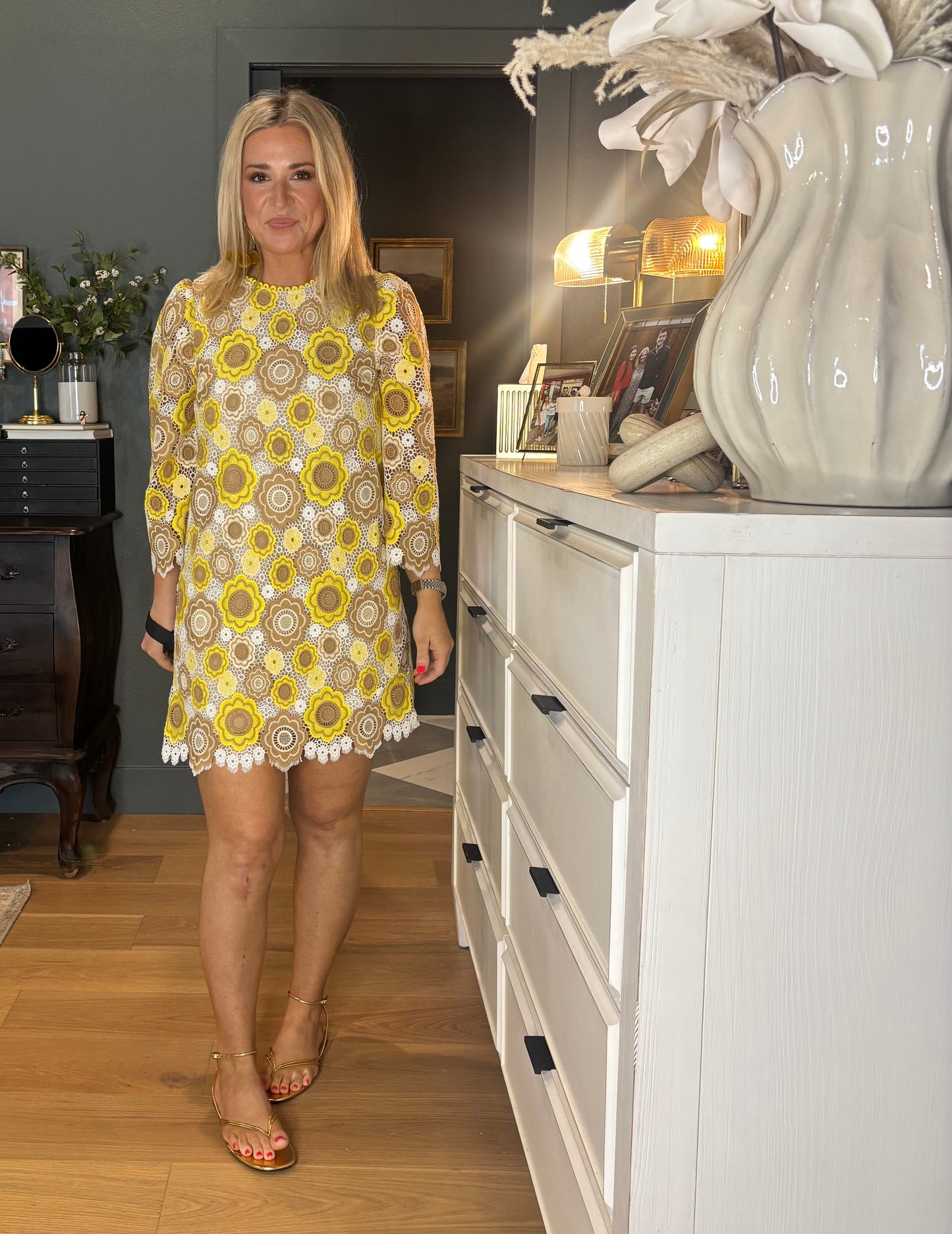 Woman in a patterned dress standing in a room with a white cabinet and decorative items.