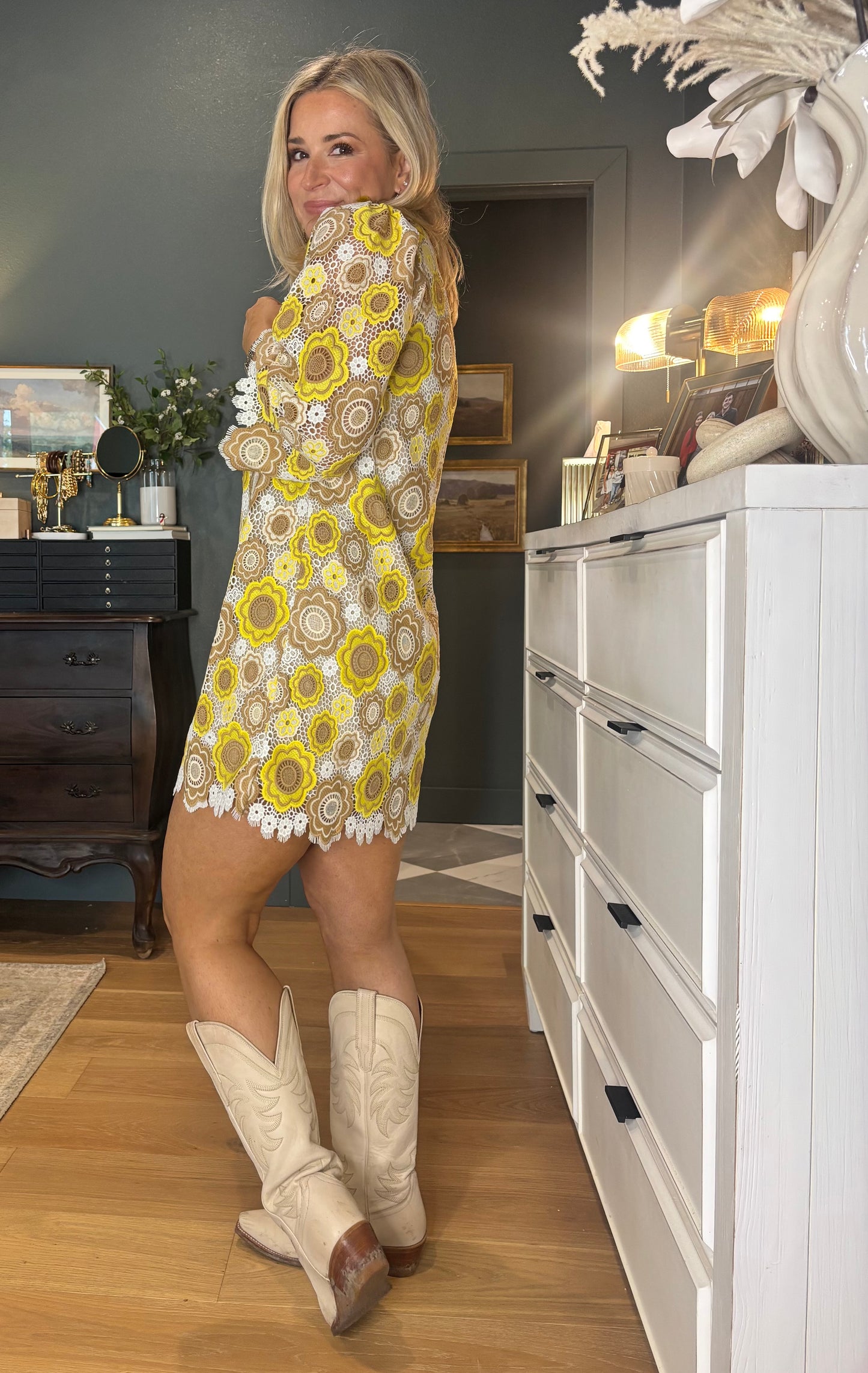 Woman in a patterned dress and white boots standing in a room with furniture and decor.
