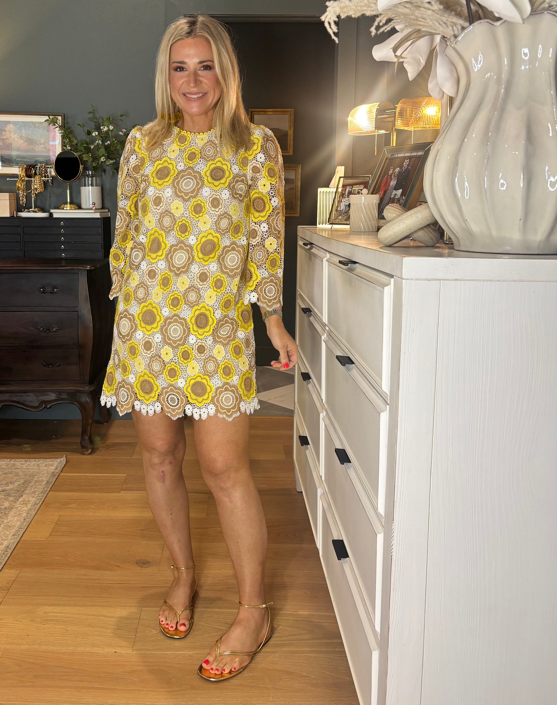 Woman in a yellow floral dress standing in a room with white cabinets and decorative items.