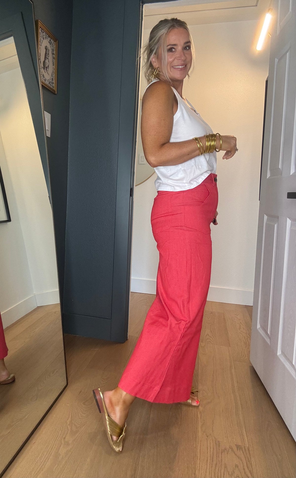 A woman posing in a white sleeveless top and bright coral red wide-leg pants, wearing gold flat sandals and bracelets, standing in a home hallway with a mirror on the left.