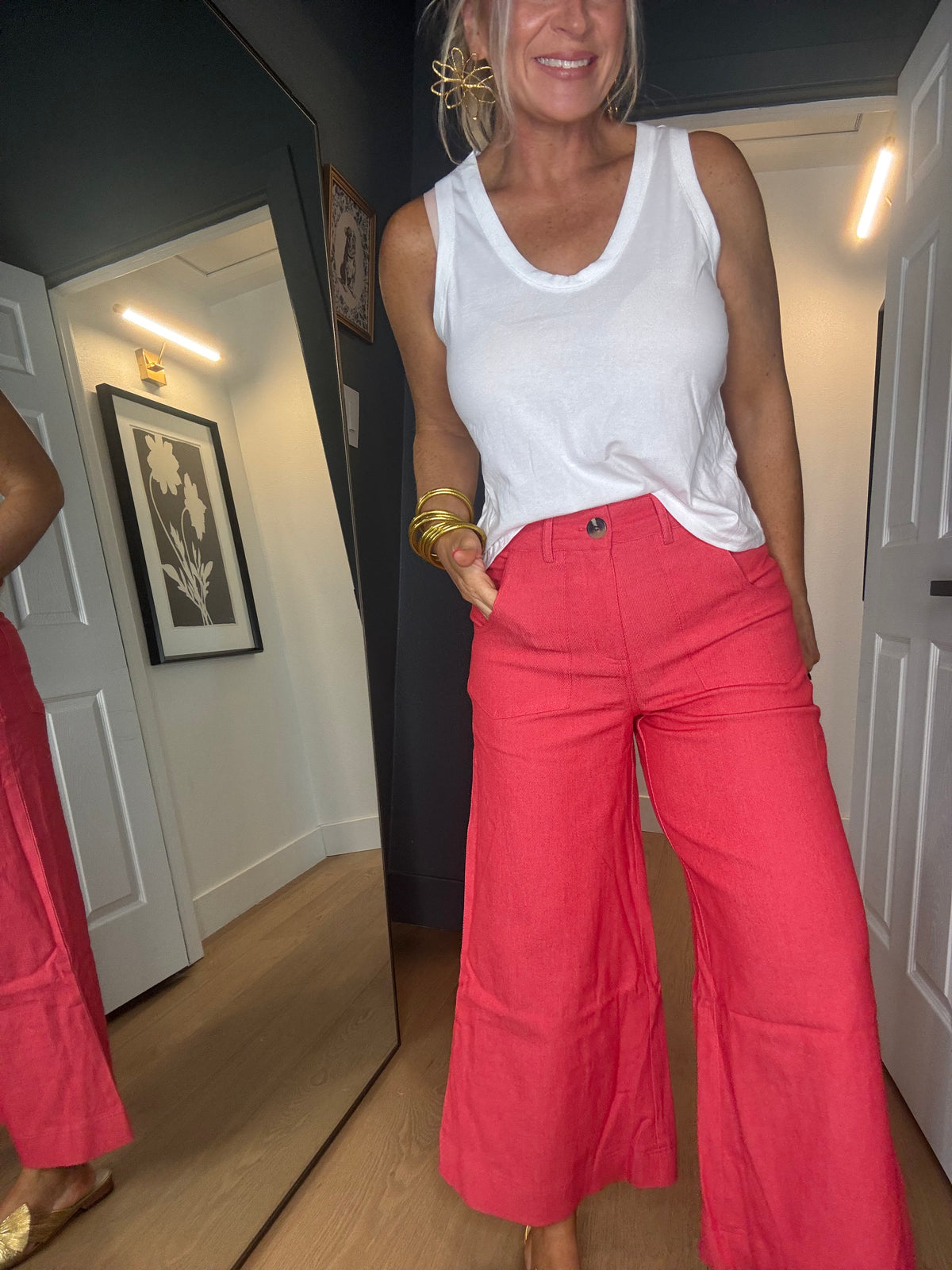 A woman wearing bright coral red wide-leg cropped pants and a white tank top, standing in a home hallway with a large mirror behind her reflecting her side.