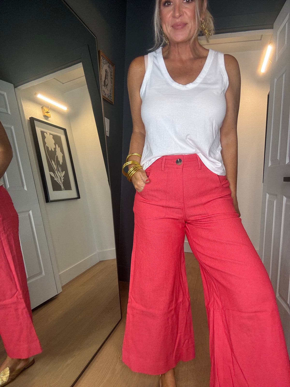A woman wearing bright red wide-leg trousers and a white tank top, standing in front of a mirror in an indoor hallway.