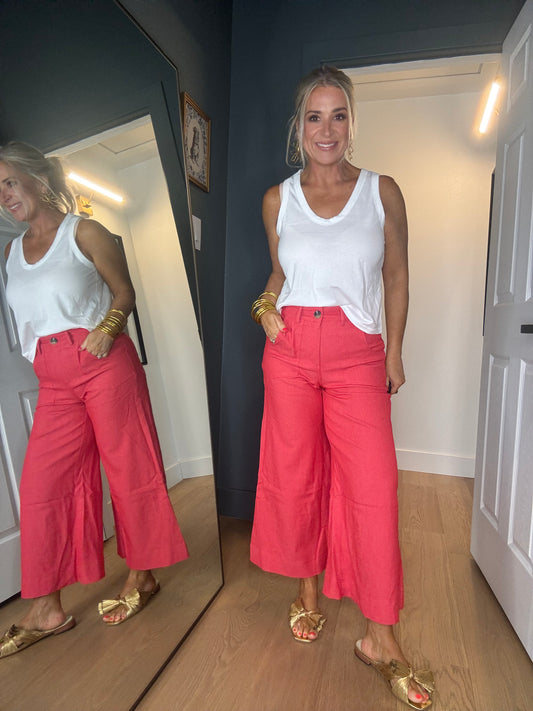 A woman modeling bright red wide-leg cropped pants with a white tank top and gold sandals, standing in a home hallway with a large mirror showing her reflection.