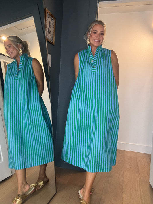 A woman wearing a sleeveless, midi-length dress with vertical green and blue stripes, standing in a room with light wood floors.