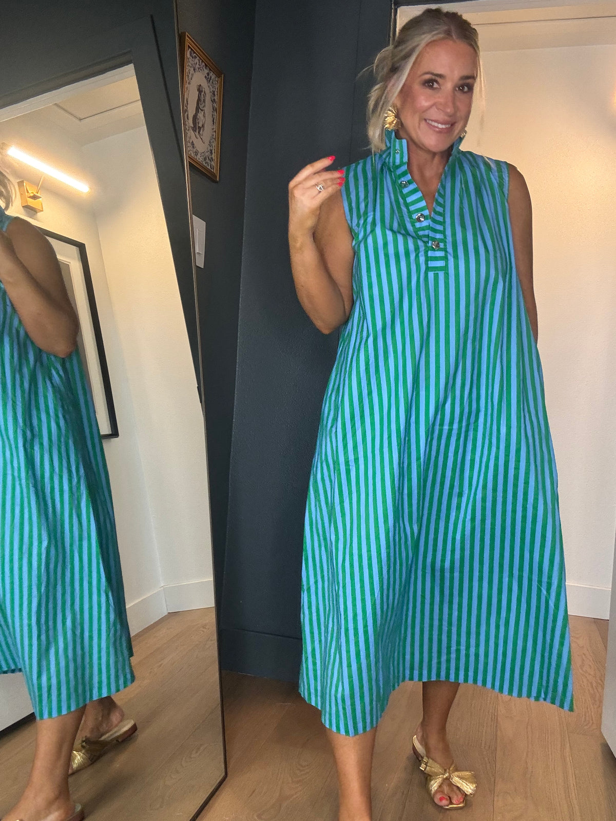 A woman wearing a sleeveless, midi-length dress with vertical green and blue stripes, standing in a room with light wood floors.