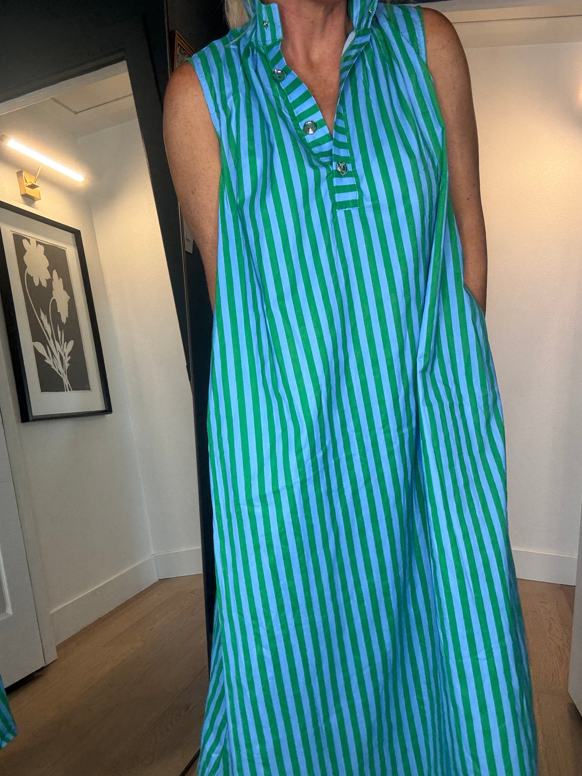 A woman wearing a sleeveless, midi-length dress with vertical green and blue stripes, standing in a room with light wood floors.