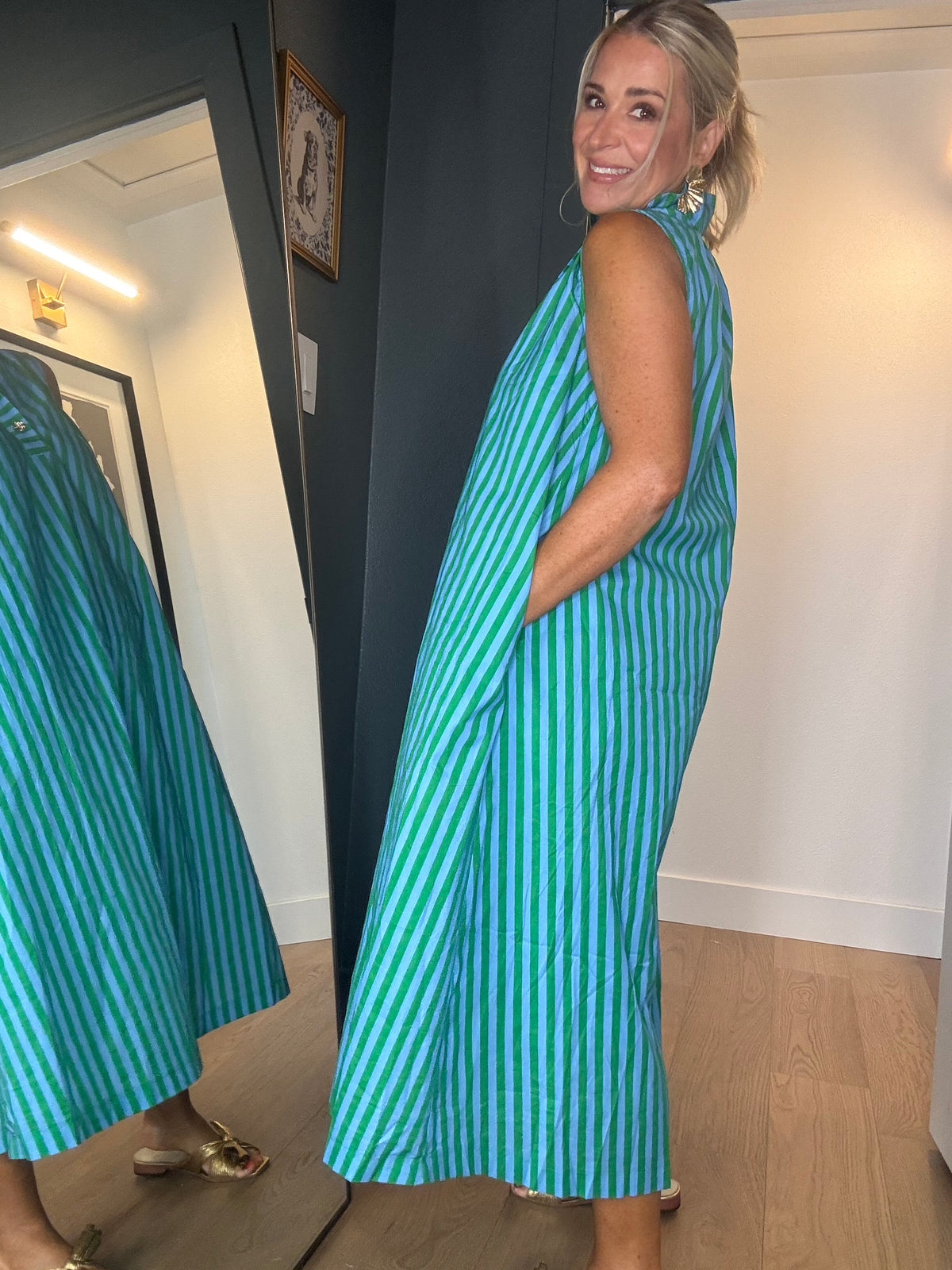 A woman wearing a sleeveless, midi-length dress with vertical green and blue stripes, standing in a room with light wood floors.
