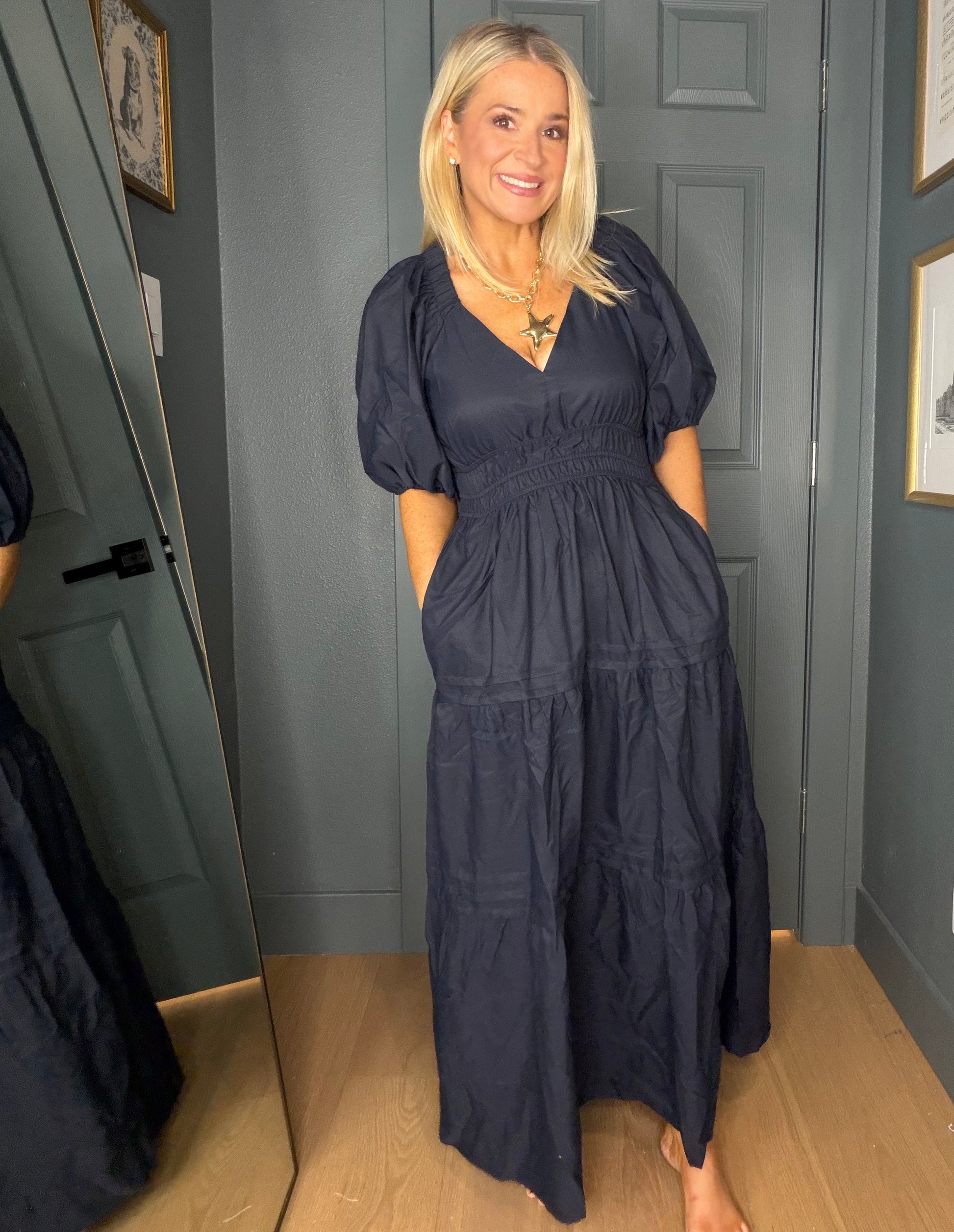 A woman wearing a dark navy blue midi dress with puff sleeves and a smocked waist, standing in front of a gray paneled door.