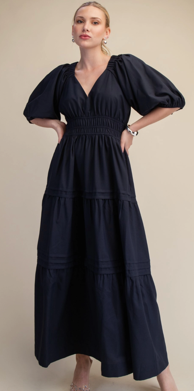 A woman modeling a navy blue midi dress with voluminous puff sleeves, a smocked waist, and a tiered skirt against a plain beige studio background.