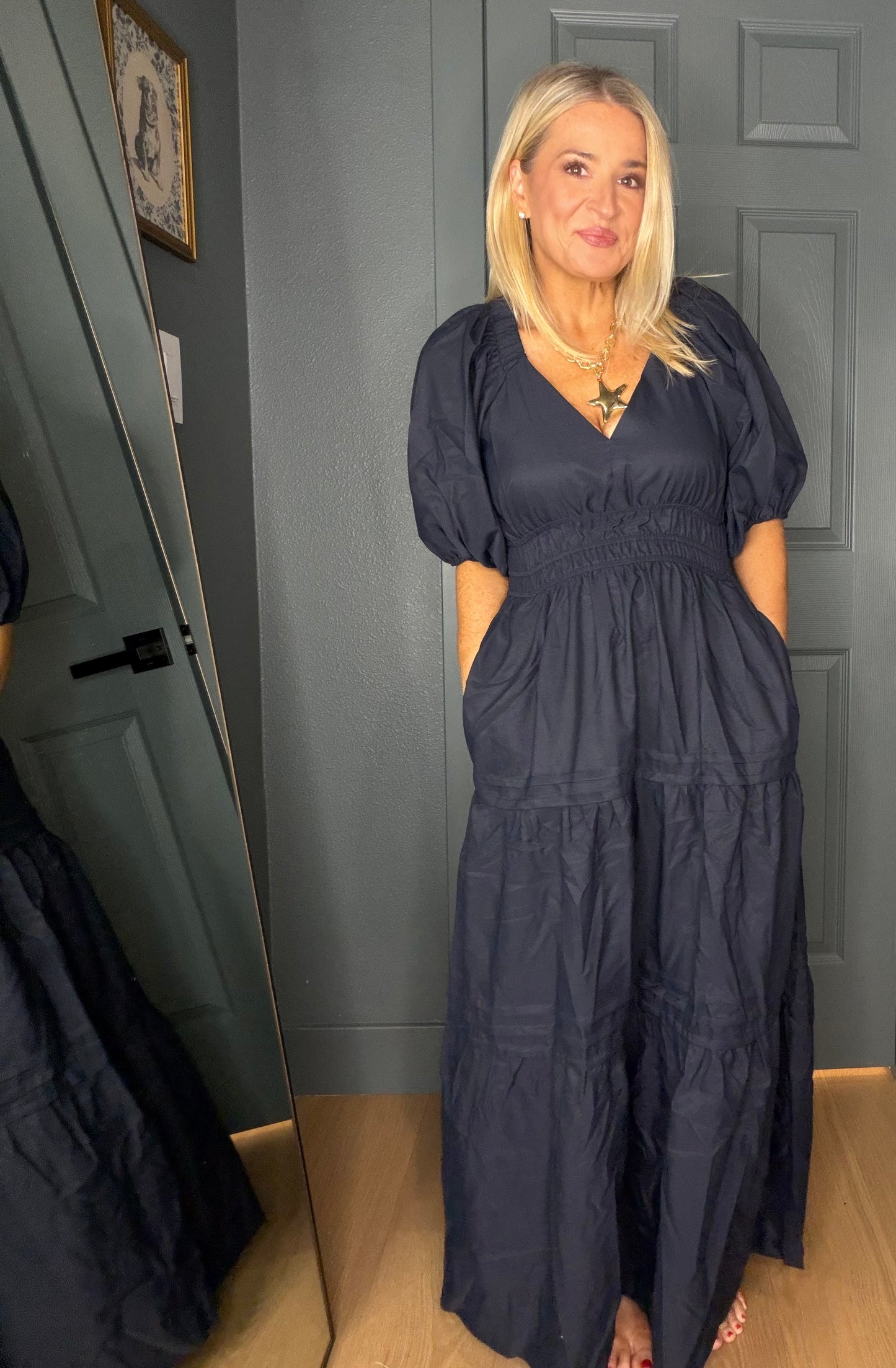 A woman wearing a dark navy blue maxi dress with puff sleeves and a smocked waist, standing in front of a gray paneled door.