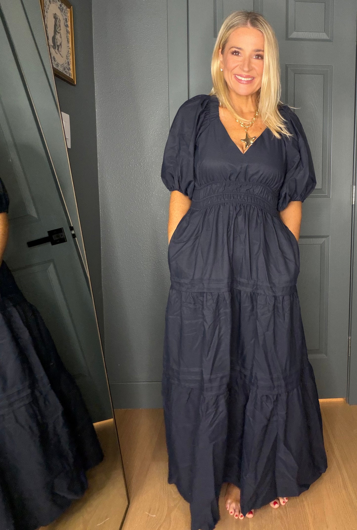 A woman modeling a navy blue maxi dress with puff sleeves, a smocked waist, and tiered skirt, standing in front of a gray paneled door.
