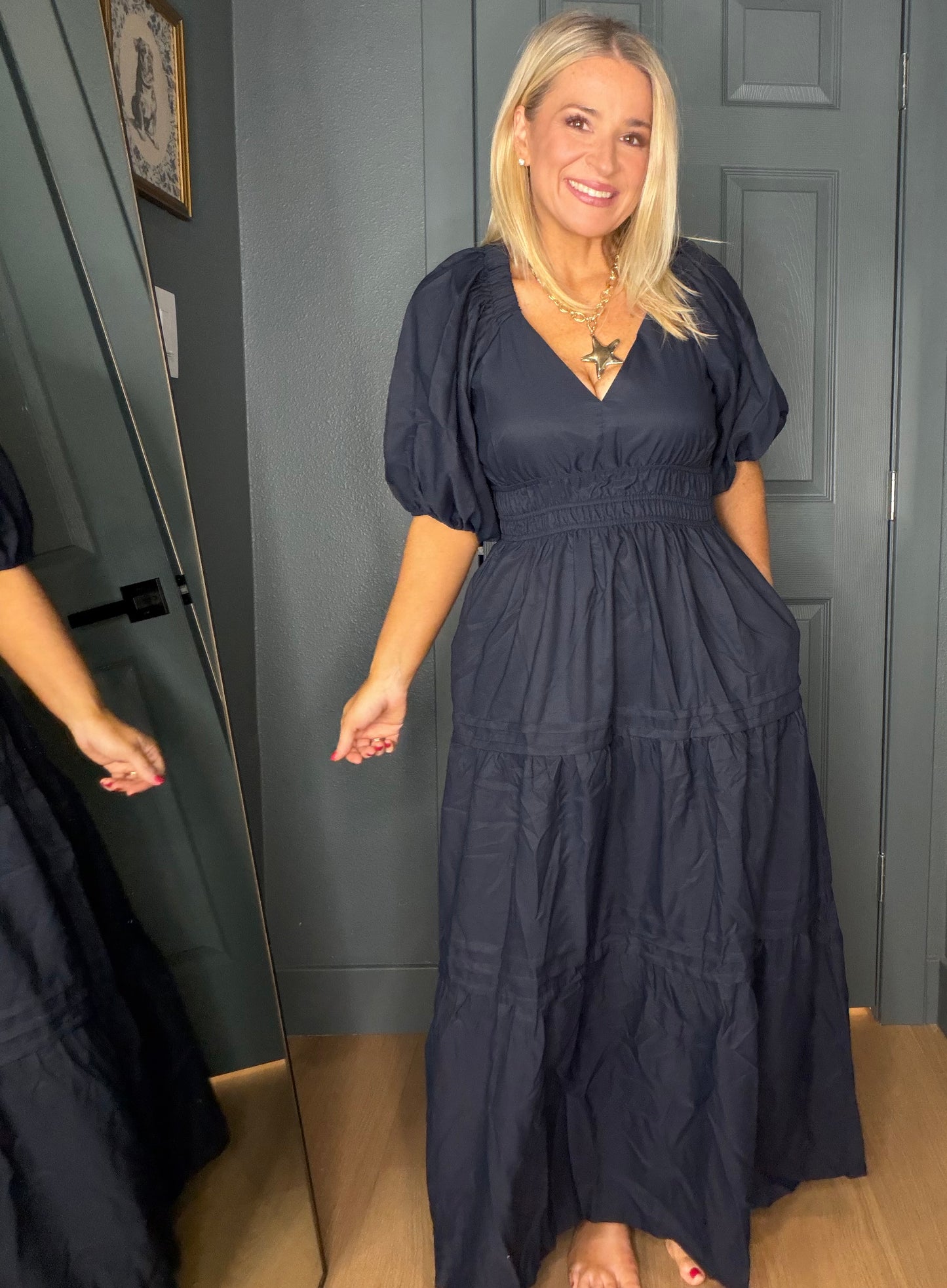 A woman wearing a dark navy blue maxi dress with puff sleeves and a smocked waist, standing in front of a gray door and mirror.