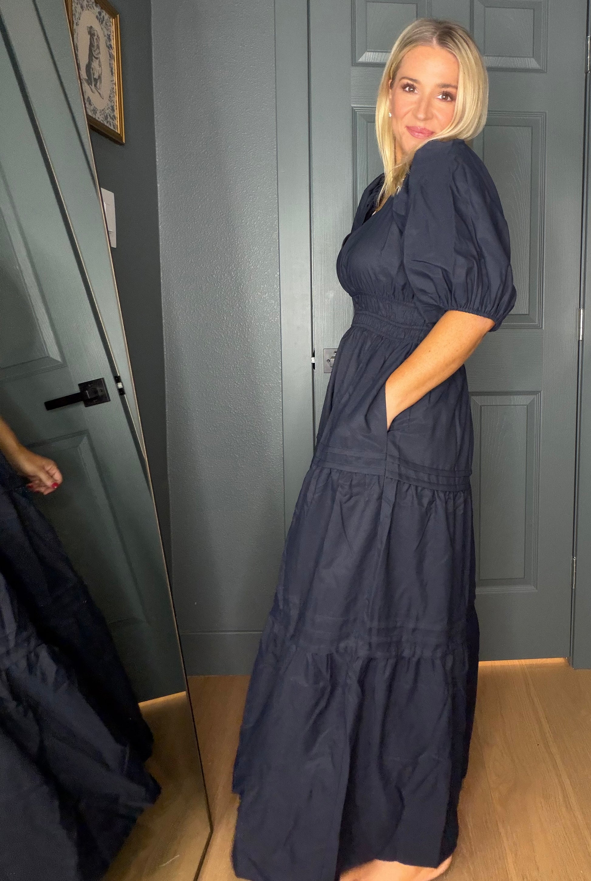 A woman wearing a dark navy blue tiered maxi dress with puff sleeves and side pockets, standing in front of gray paneled doors.