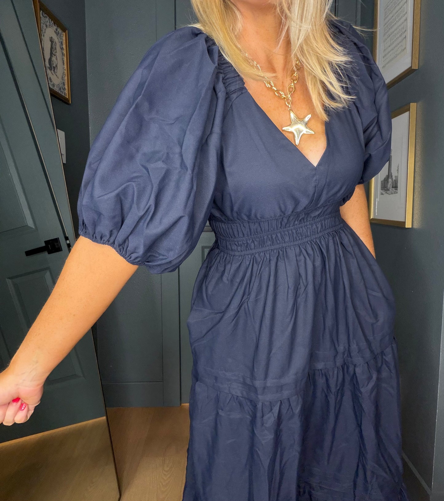 A woman wearing a navy blue tiered dress with puff sleeves and a smocked waist, standing in an indoor room with dark gray walls and framed pictures.