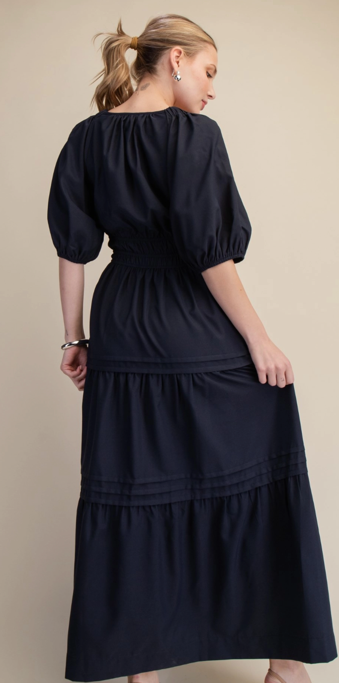 A woman wearing a dark navy midi dress with puff sleeves, a smocked waist, and a tiered skirt, shown from the back against a solid beige background.