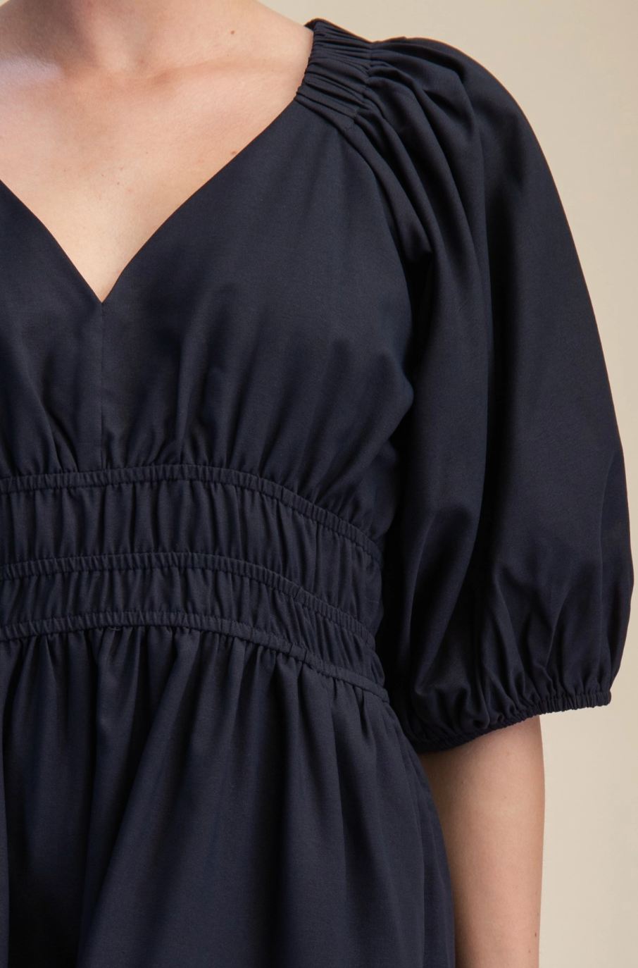 Close-up of a person wearing a dark navy dress with a V-neckline, gathered puff sleeves, and an elasticized waist seam against a plain beige background.