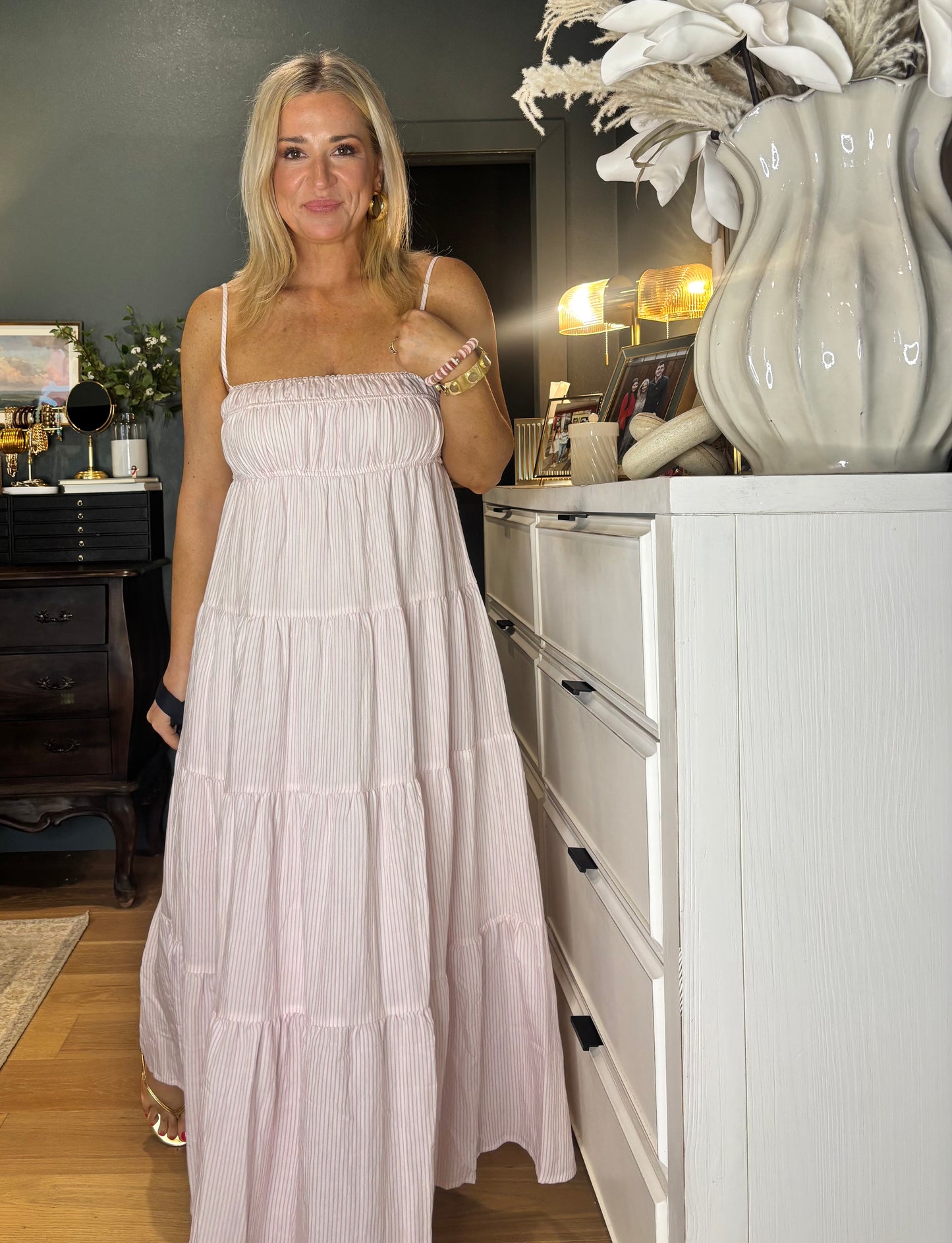 Woman in a long, light-colored dress standing in a room with a white dresser and decorative items.