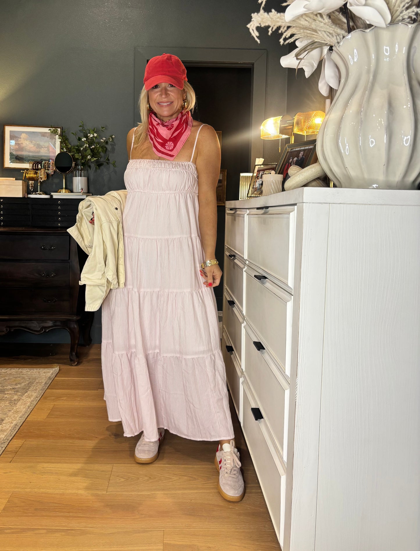 Woman in a long light pink dress standing in a room with a white dresser and decorative items.