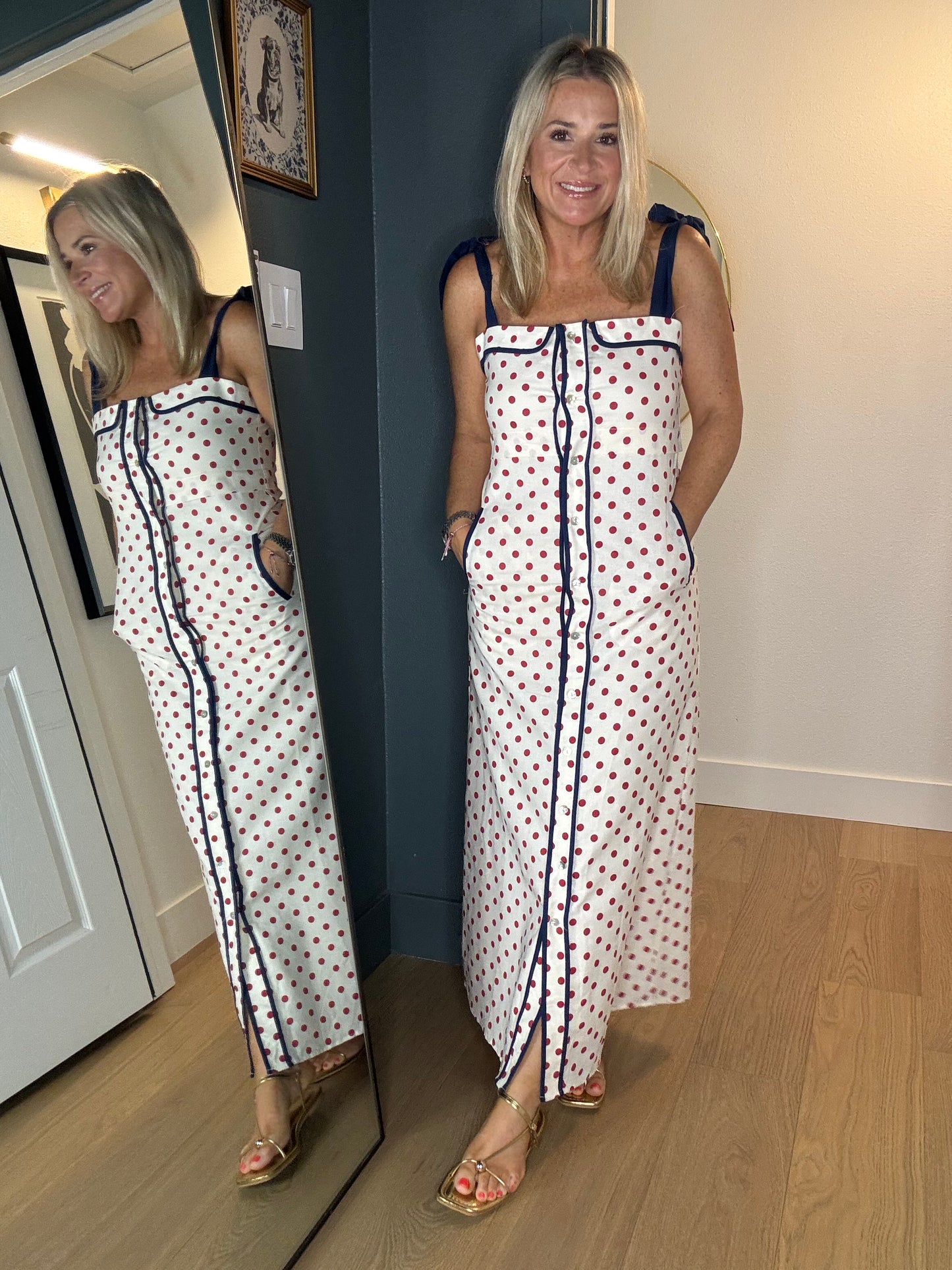 A woman wearing a white maxi dress with red polka dots and navy blue trim, standing in an indoor room with a wooden floor and a mirror showing her reflection.