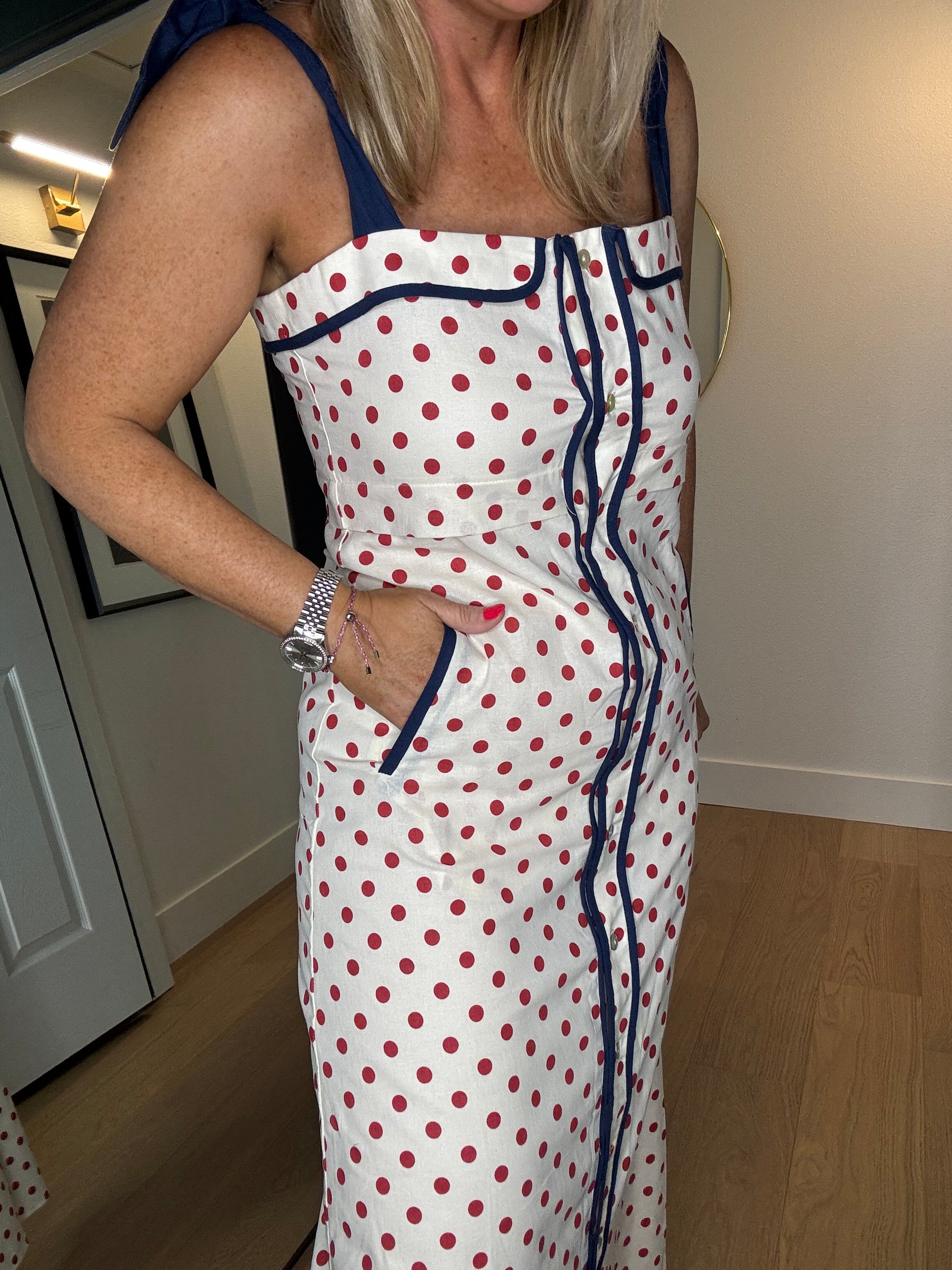A woman wearing a white sleeveless dress with red polka dots and navy blue trim, photographed indoors in a home setting.