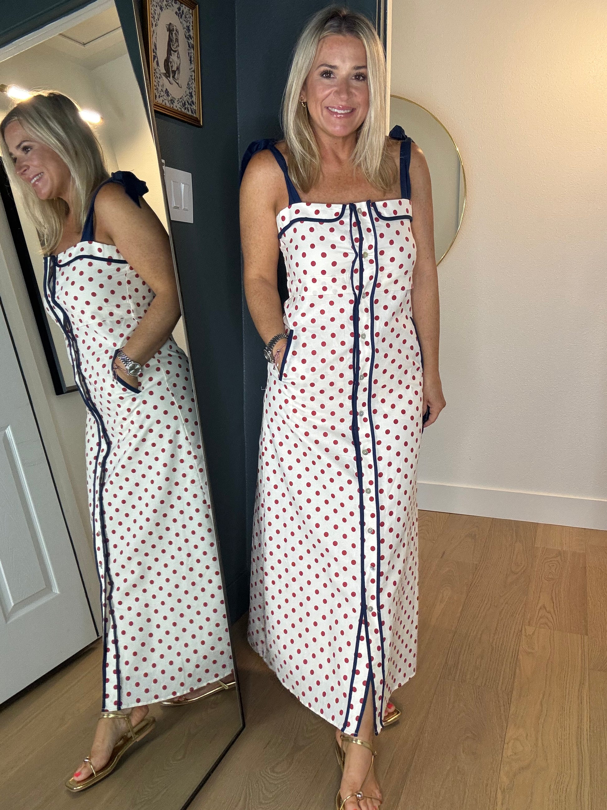 A woman modeling a white sleeveless maxi dress with red polka dots and navy blue trim, standing in front of a mirror that reflects her side profile. The room has light wood flooring and a dark gray accent wall.