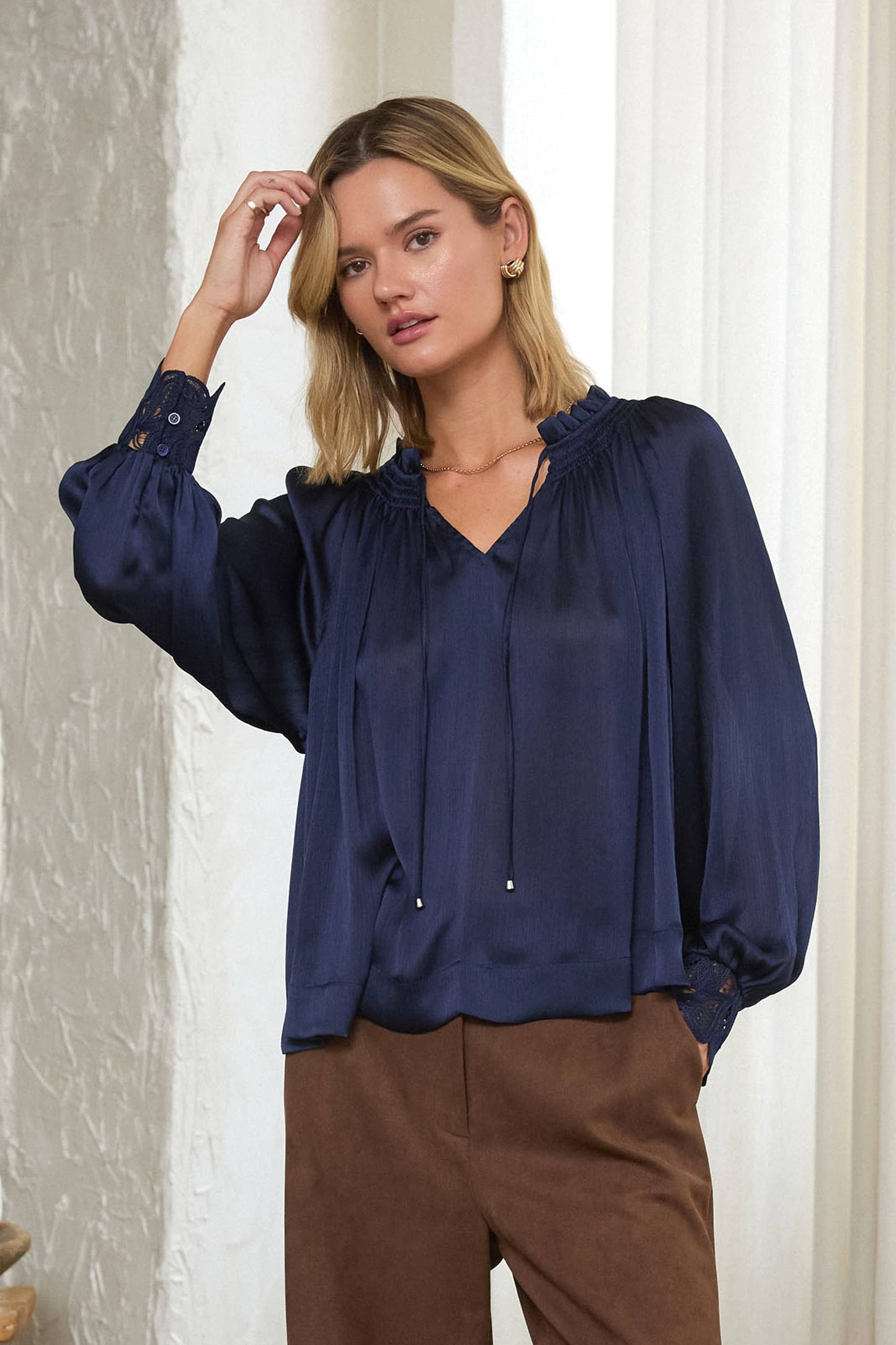 A woman wearing a navy blue long-sleeve blouse with a gathered ruffled neckline and brown trousers, posing indoors against a textured white wall and white curtain.