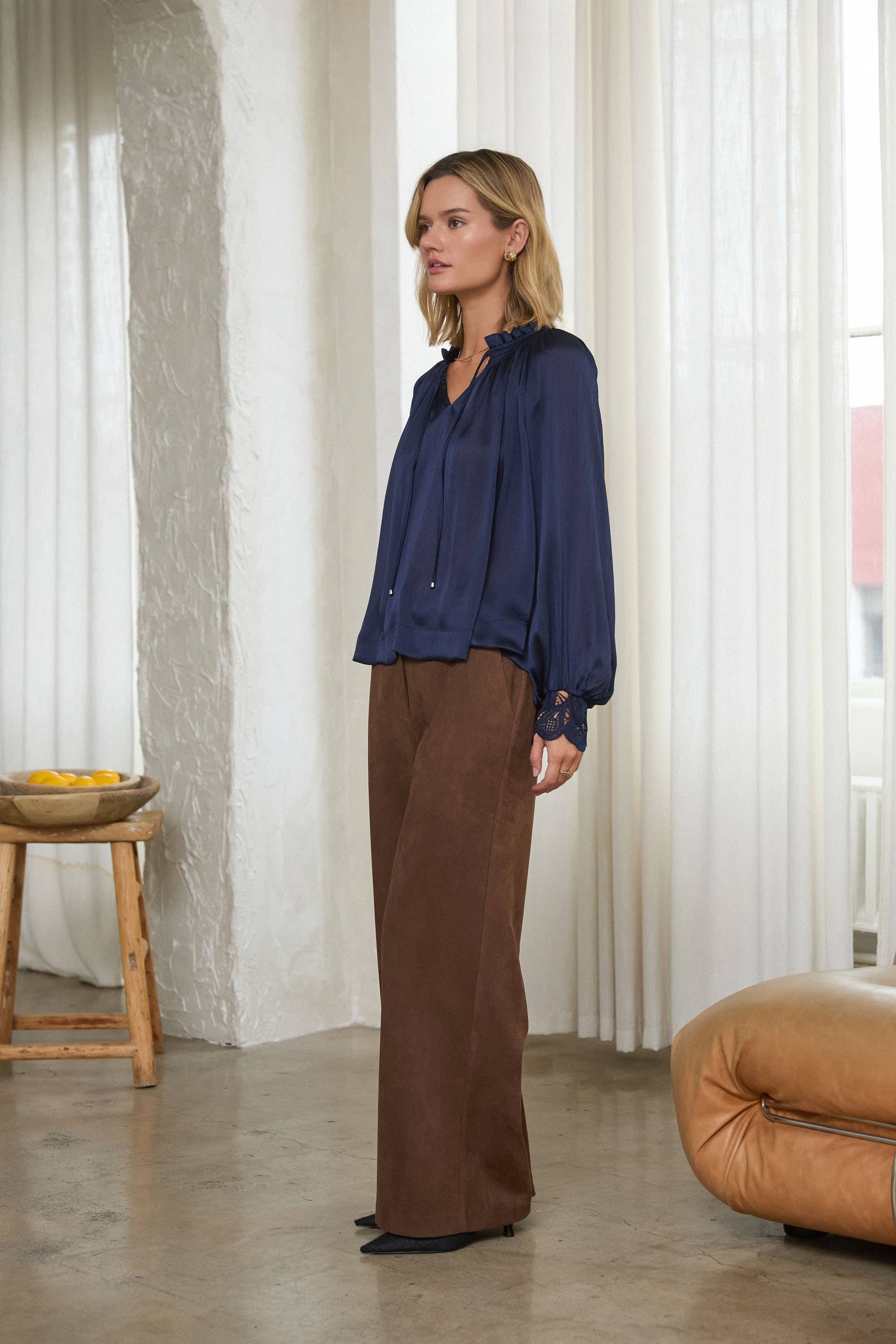 A woman stands in a bright indoor room wearing a navy blue long-sleeve blouse and brown wide-leg trousers, with white curtains, a small wooden stool, and a tan pouf in the background.
