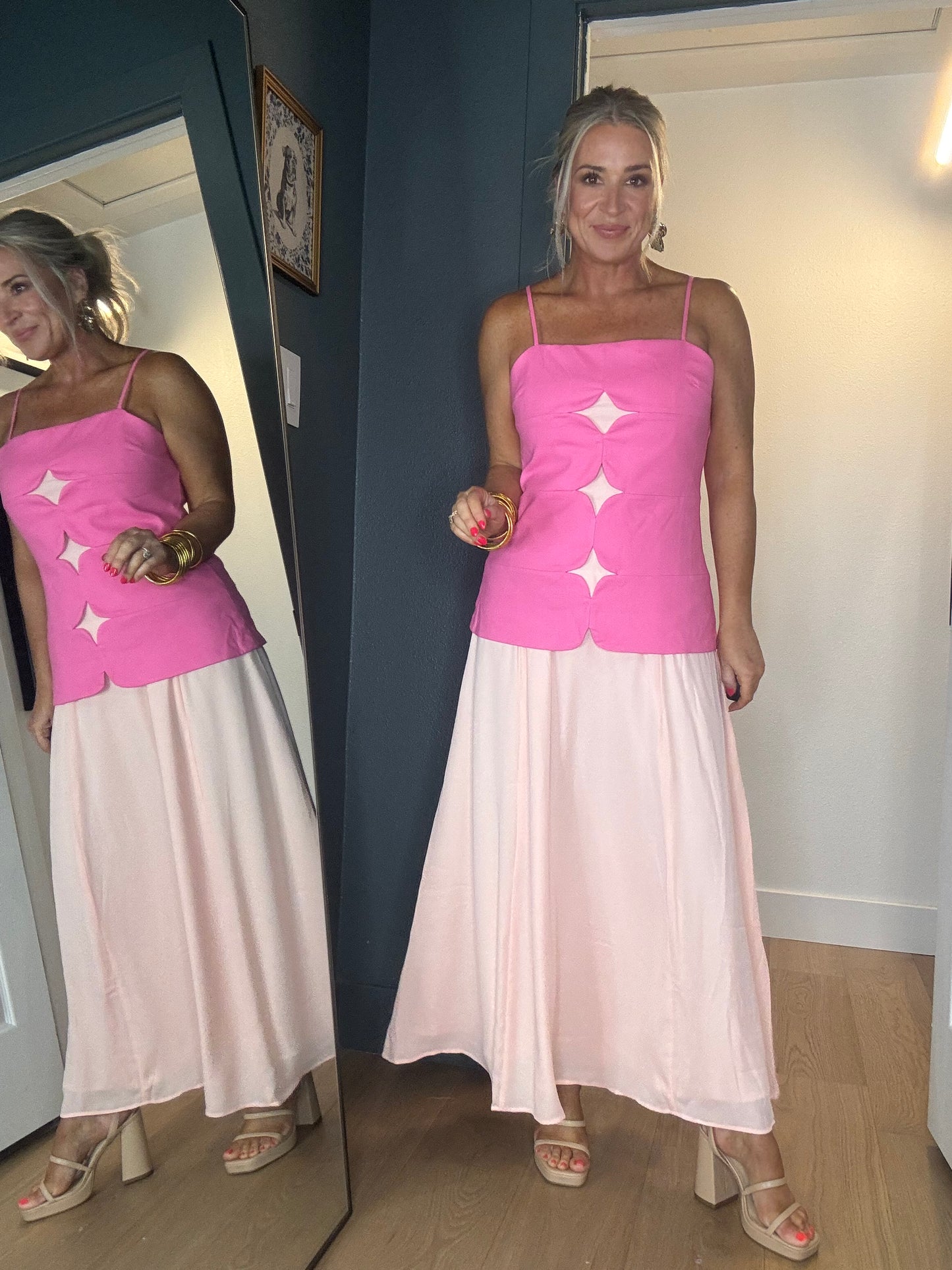 A woman wearing a bright pink sleeveless midi dress with thin straps and three large mesh star-shaped cutouts down the front, paired with a long light peach skirt.