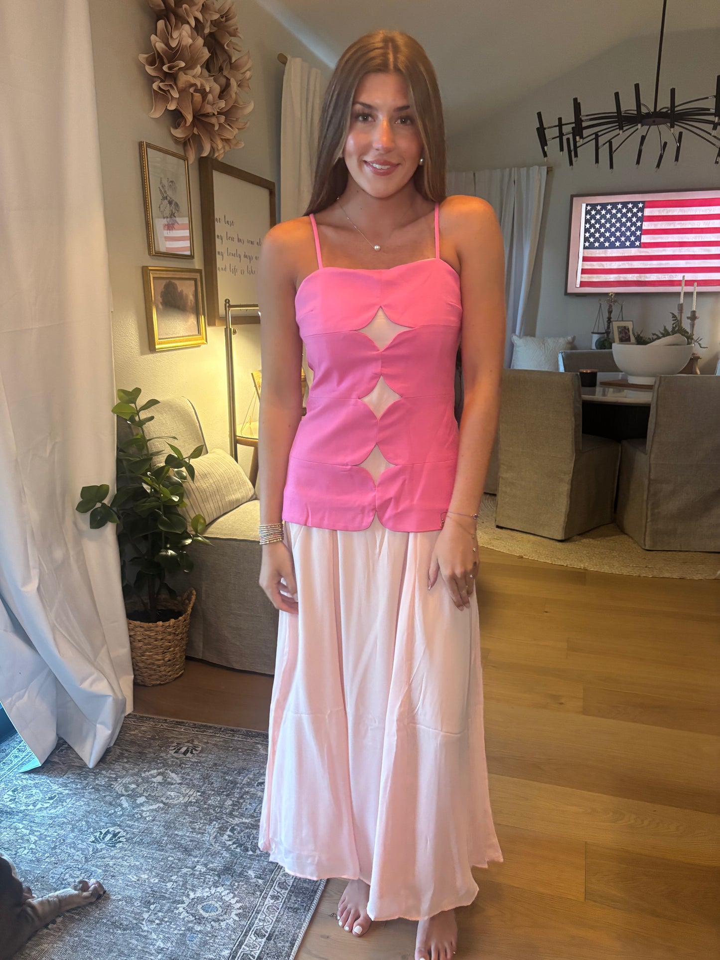 A woman stands indoors wearing a bright pink sleeveless camisole top with diamond-shaped cutouts and a long, flowing light pink skirt. The photo is taken in a home interior with wood floors, a rug, curtains, and furniture visible in the background.