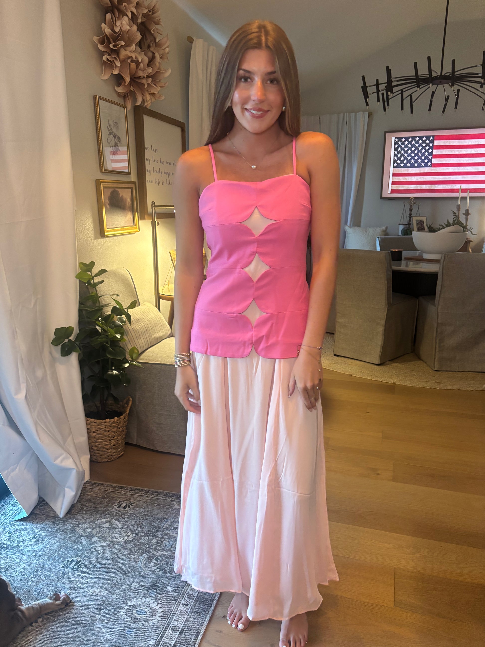 A woman stands indoors wearing a bright pink sleeveless camisole top with diamond-shaped cutouts and a long, flowing light pink skirt. The photo is taken in a home interior with wood floors, a rug, curtains, and furniture visible in the background.