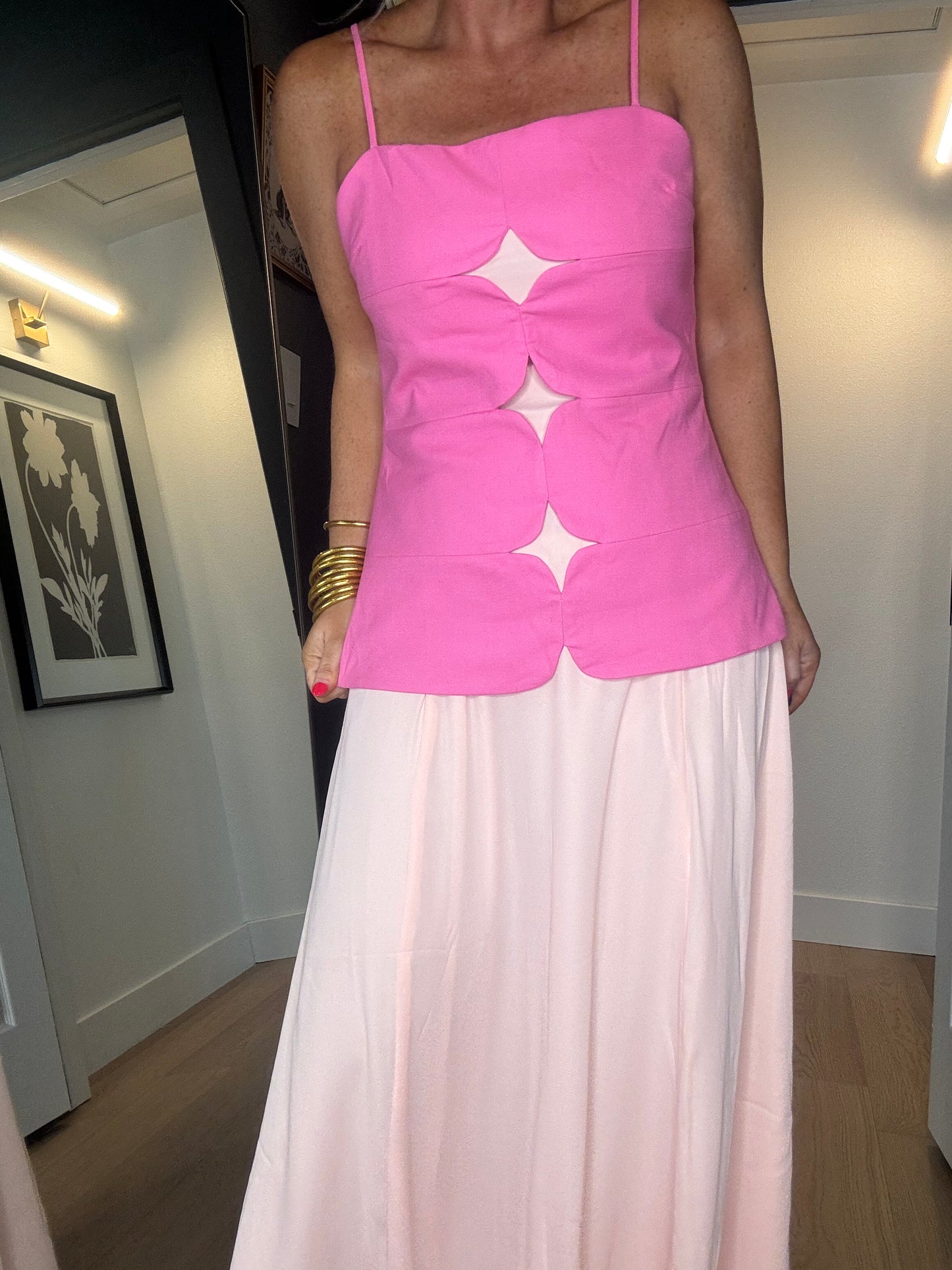 A woman wearing a bright pink sleeveless midi dress with thin straps and three large mesh star-shaped cutouts down the front, paired with a long light peach skirt.