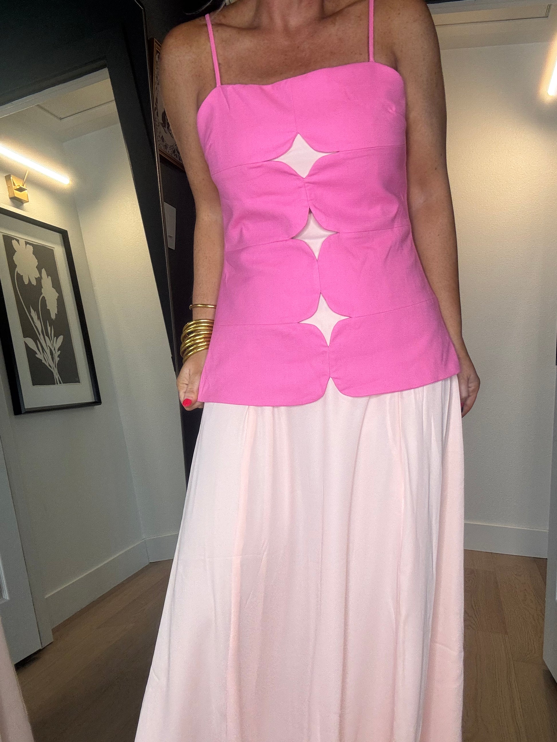 A woman wearing a bright pink sleeveless midi dress with thin straps and three large mesh star-shaped cutouts down the front, paired with a long light peach skirt.