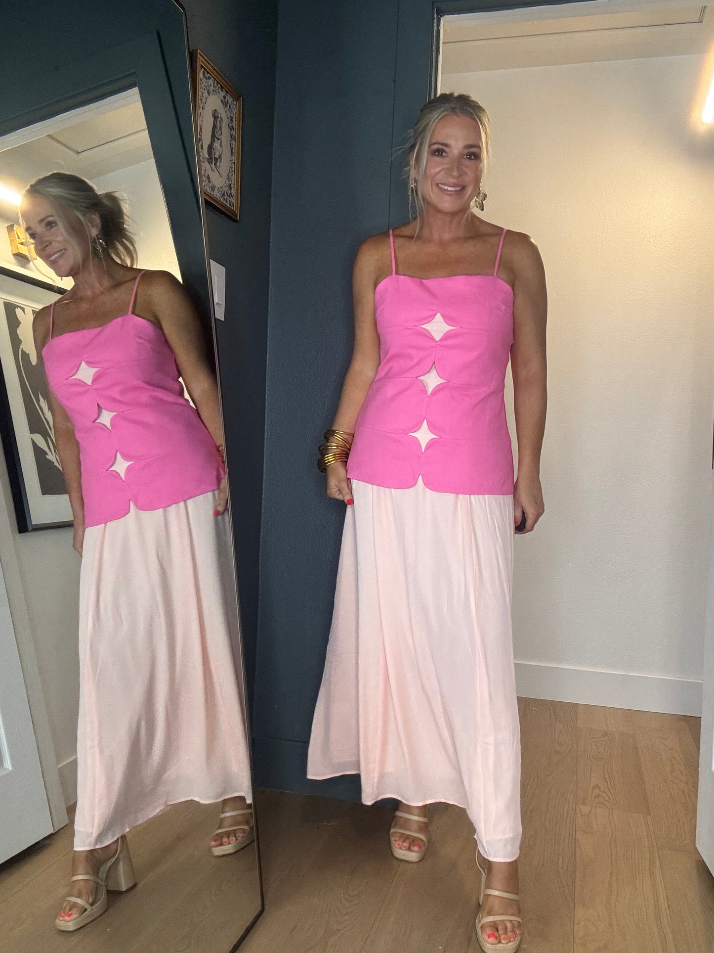A woman wearing a bright pink sleeveless midi dress with thin straps and three large mesh star-shaped cutouts down the front, paired with a long light peach skirt.