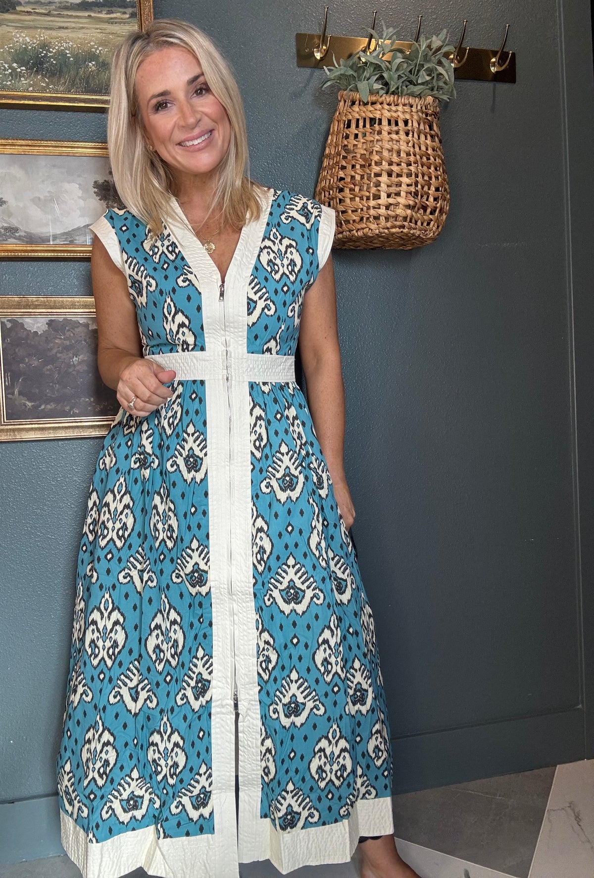 A woman modeling a sleeveless V-neck maxi dress with a blue, white, and black ikat pattern, standing against a dark gray wall with framed art and a hanging basket.