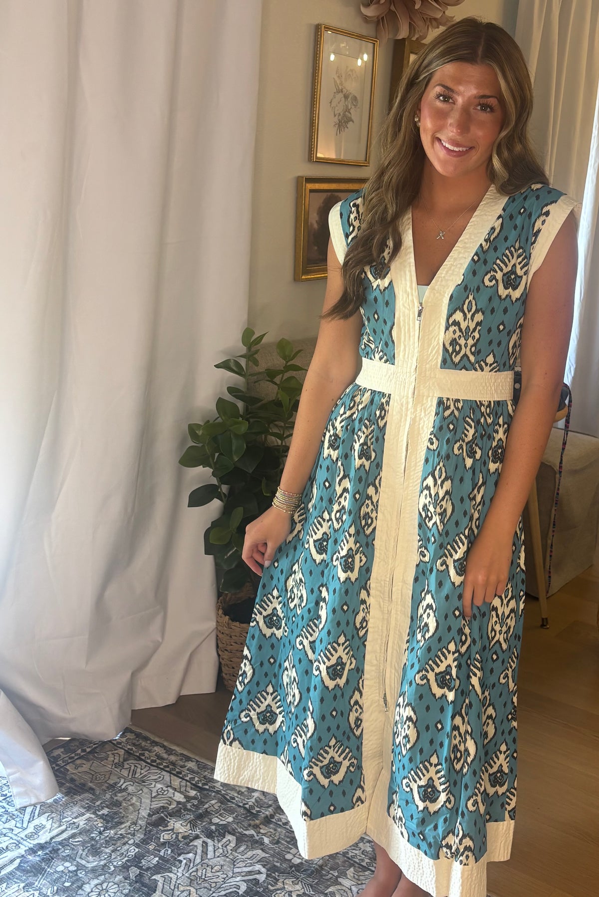 A woman wearing a sleeveless V-neck midi dress with a teal and cream ikat pattern, standing in a home setting with a white curtain and a potted plant in the background.