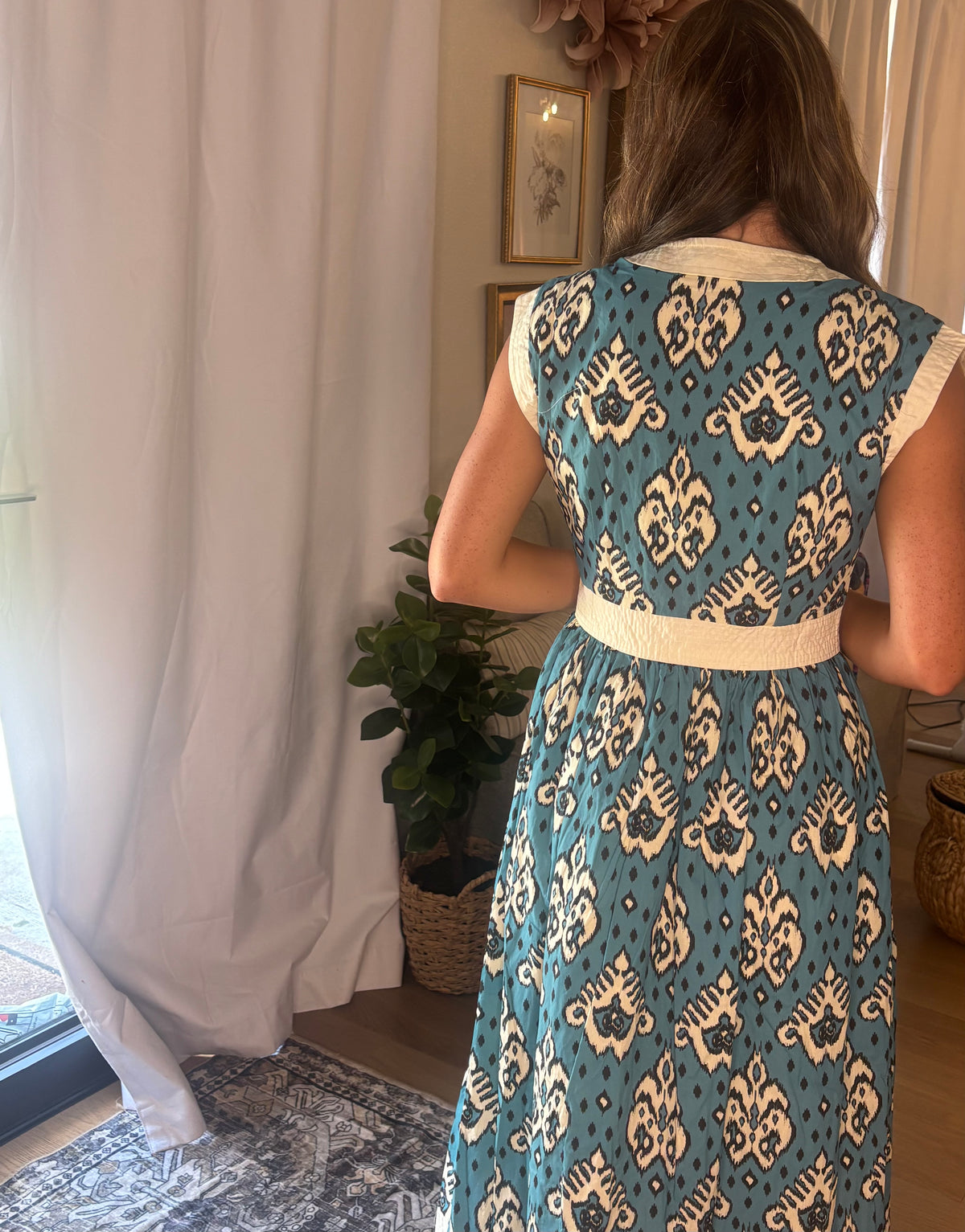 A woman viewed from behind wearing a sleeveless midi dress with a teal blue and cream ikat pattern, standing in a room with white curtains and a potted plant.