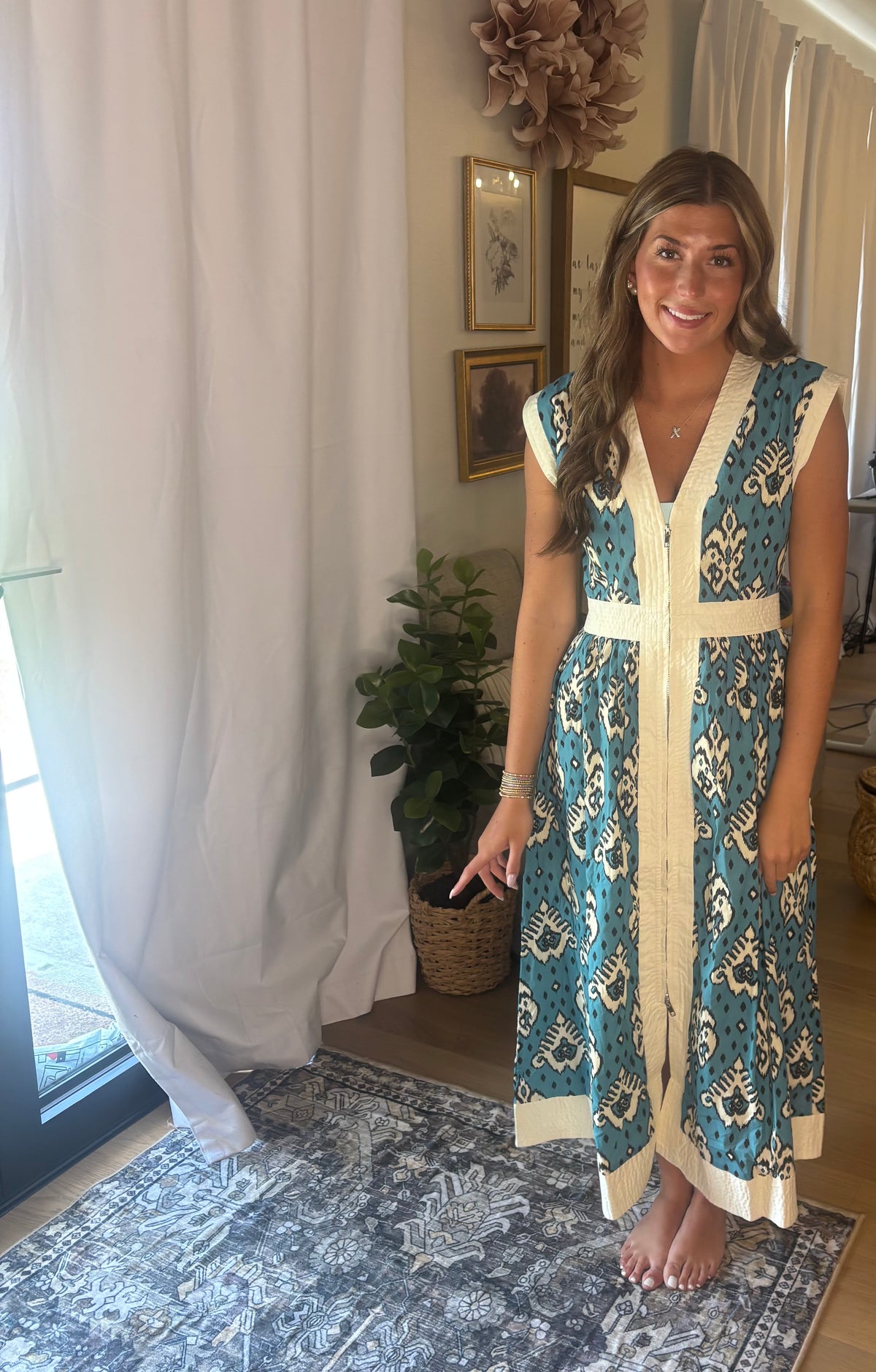 A woman modeling a sleeveless teal and cream bohemian-style maxi dress in a home setting with a patterned rug, white curtains, and a potted plant.