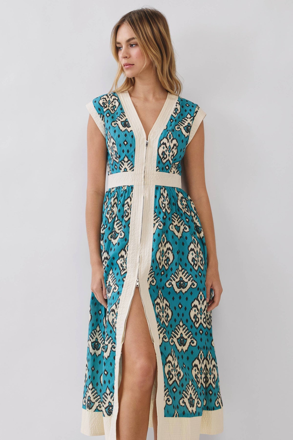 A woman modeling a sleeveless, V-neck maxi cover-up dress with a teal blue and cream ikat pattern, set against a plain light gray background.