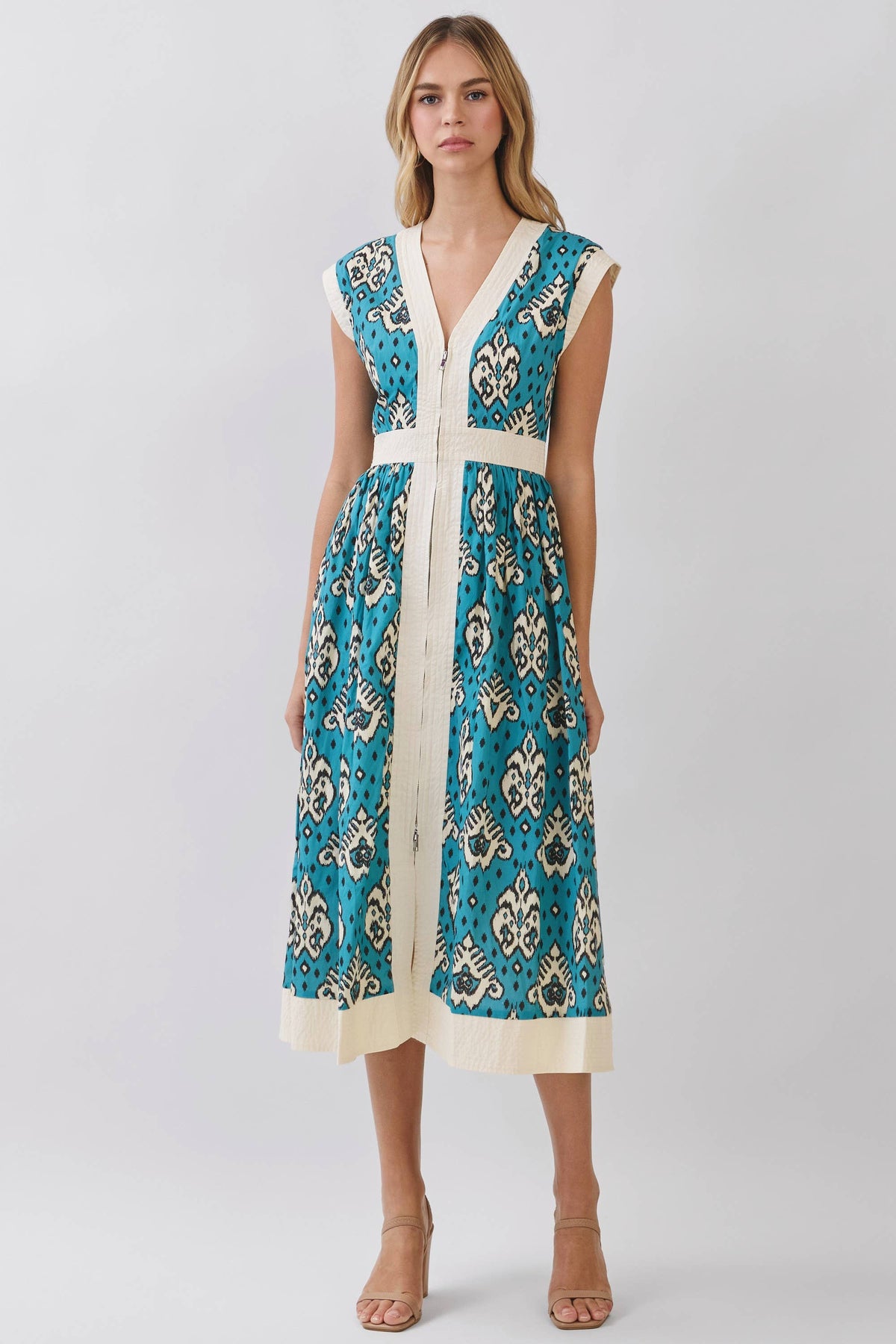 A woman modeling a sleeveless midi dress with a teal blue and cream paisley pattern, featuring a cream V-neckline, waistband, and side panels, set against a plain light gray studio background.