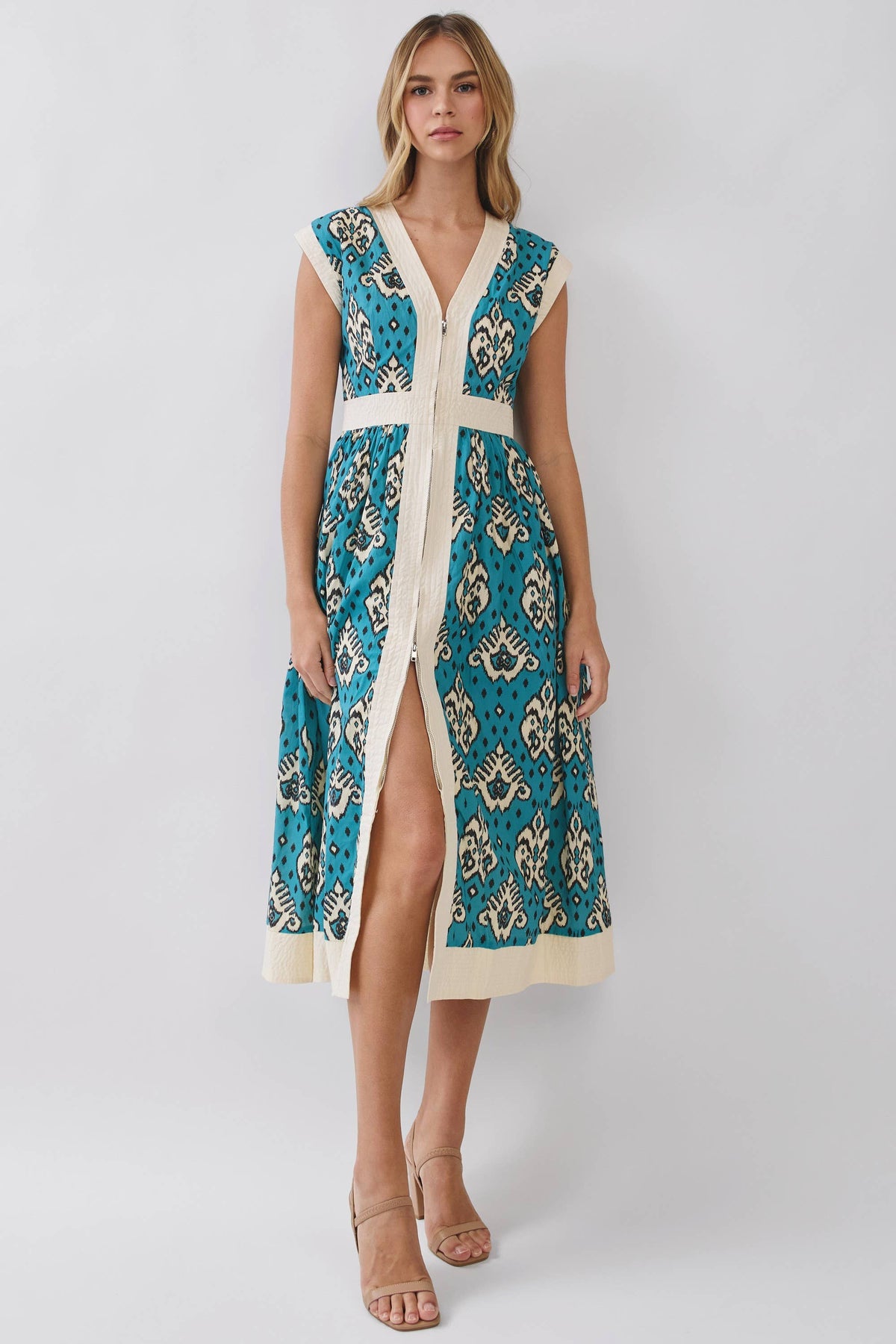 A woman modeling a sleeveless midi dress with a teal and cream paisley pattern, featuring a V-neckline, front button closure, and a high side slit, set against a plain light gray studio background.