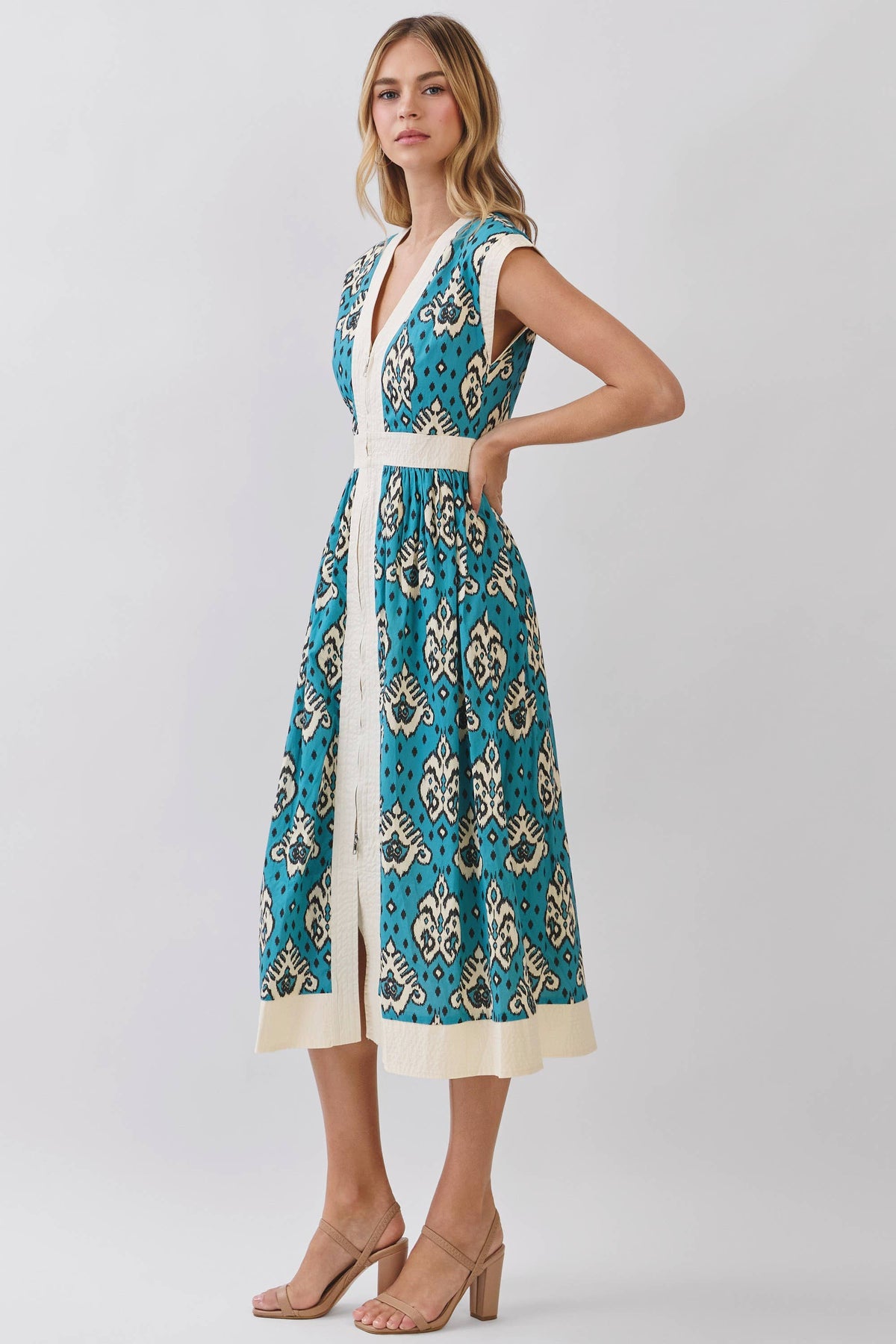 A woman modeling a sleeveless midi dress with a teal blue and cream paisley pattern, featuring a white V-neckline, white waistband, and a cream hem, set against a plain light gray studio background.