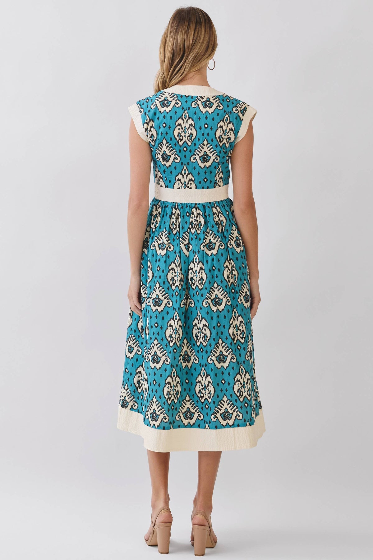 A woman viewed from the back wearing a sleeveless midi dress with a turquoise blue and cream patterned print, a cream waistband, and a cream hem, paired with beige high-heeled sandals against a light gray studio background.